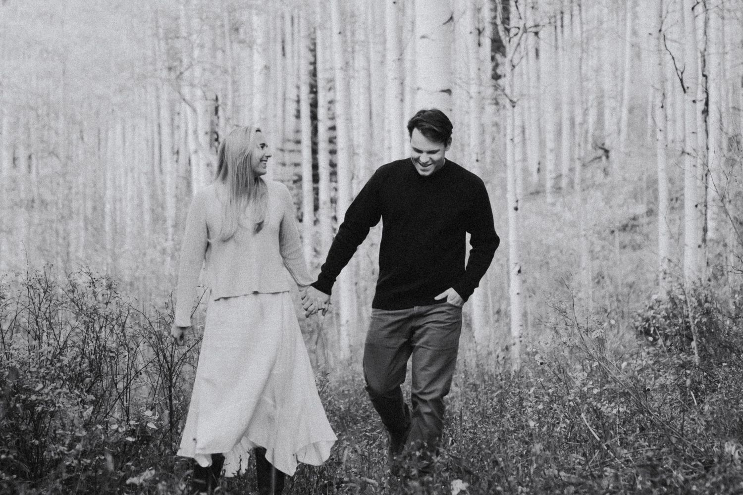 A couple walks hand-in-hand through an enchanting aspen forest, smiling at each other, showcasing their love and connect