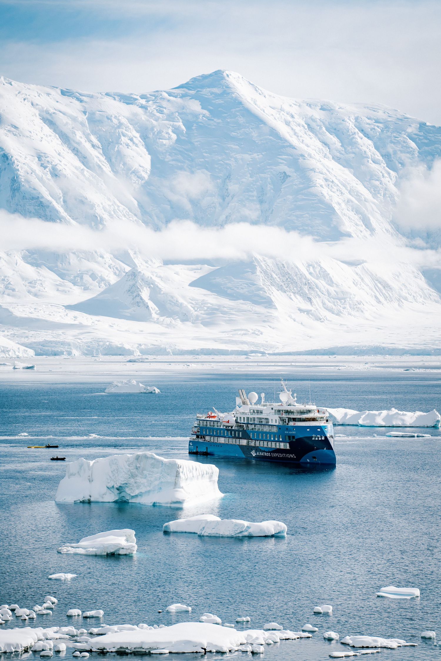 A blue cruise ship navigates through ice-filled Antarctic waters with snow-covered mountains in the background.