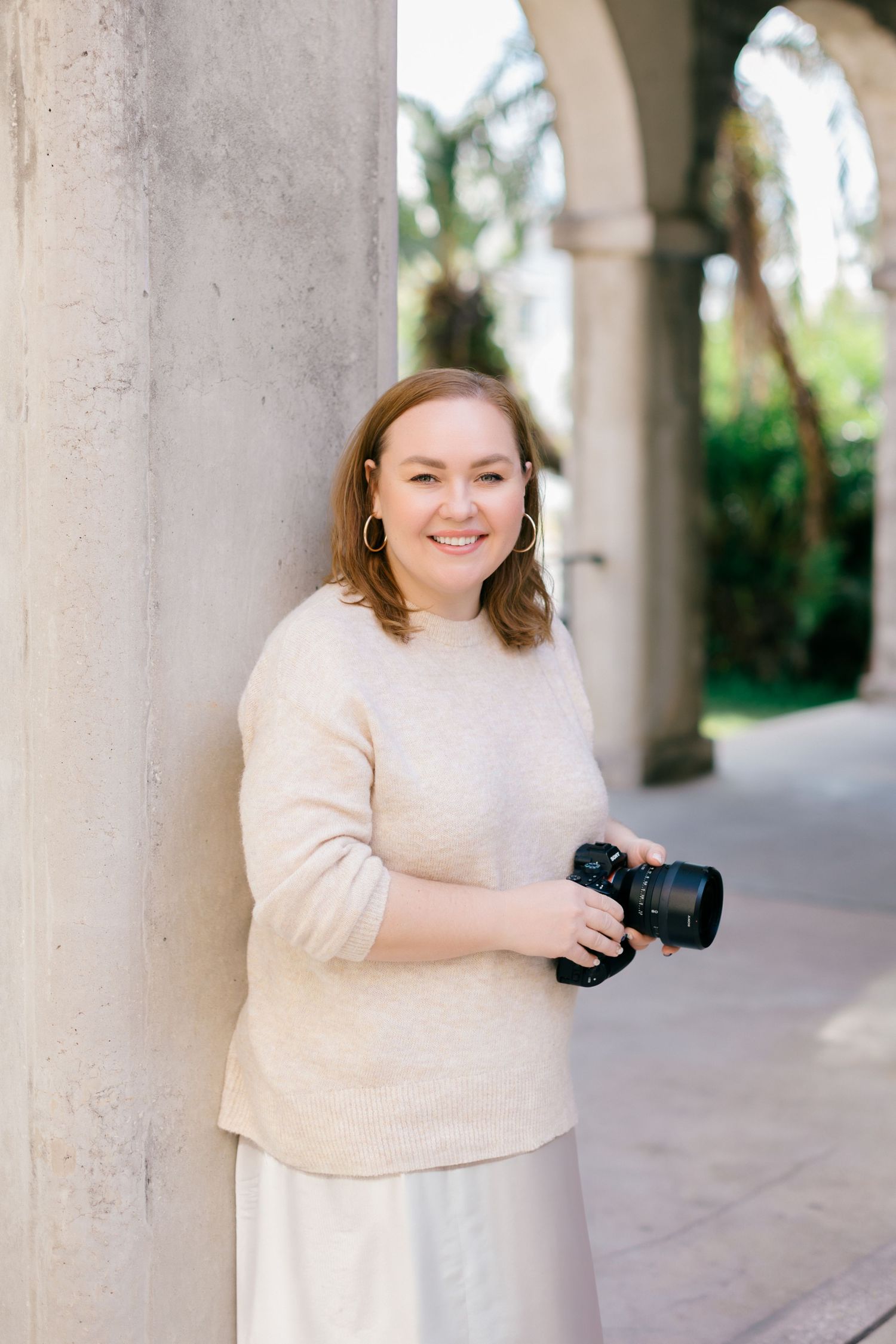 Professional photographer in cream sweater poses with camera in columned archway.
