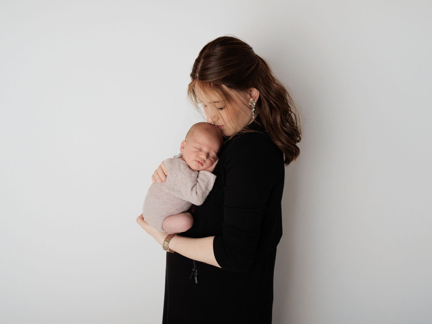 A tender moment between parent and newborn baby against a white wall, with the adult wearing a black sweater.