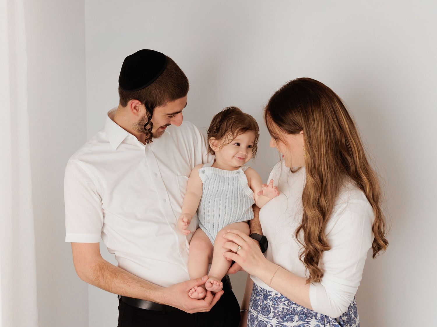 A young Jewish family with a man, woman, and baby sharing a tender moment against a white wall.