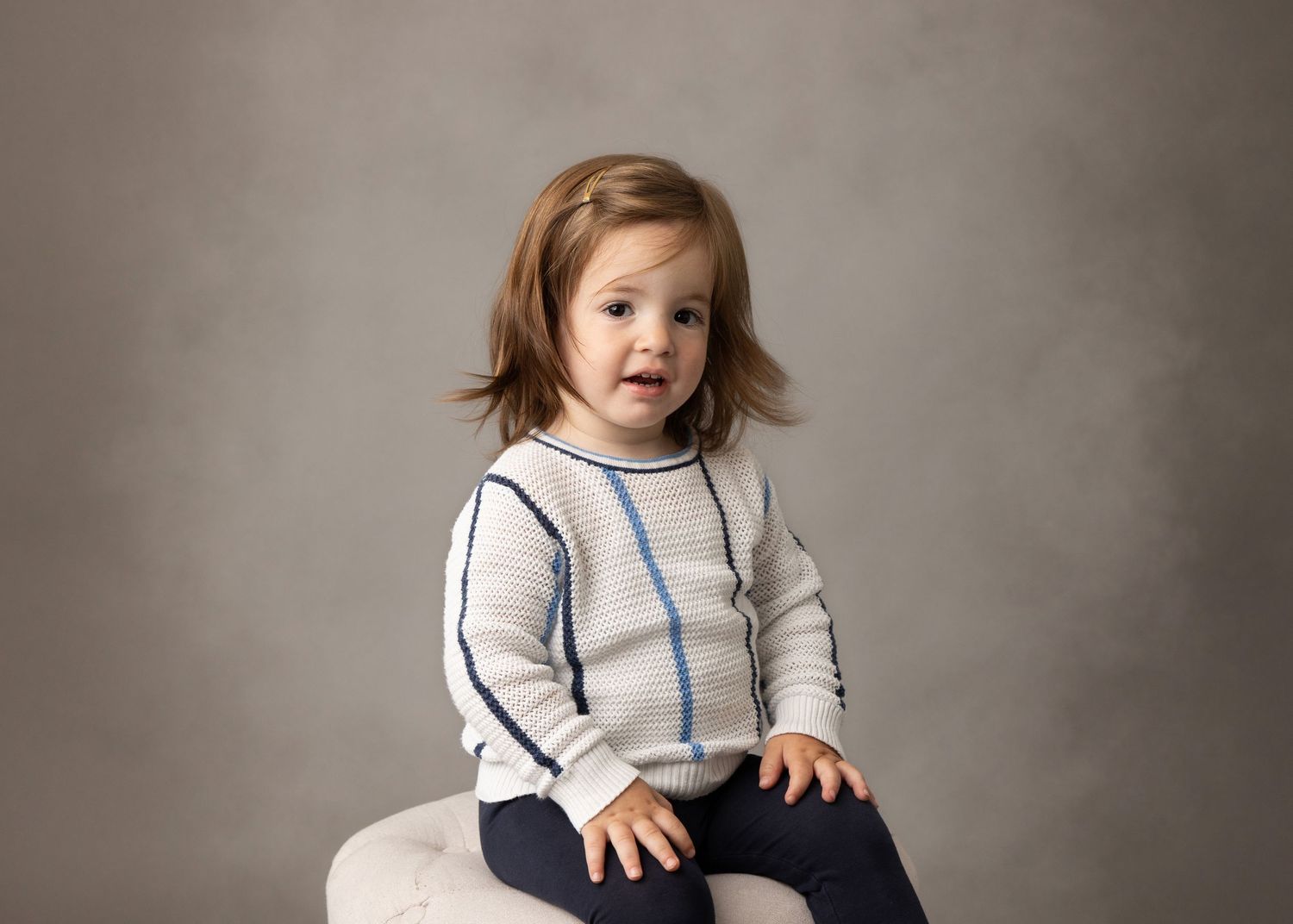 Baby wearing white knit sweater sitting on beige backdrop with a playful expression during portrait session.