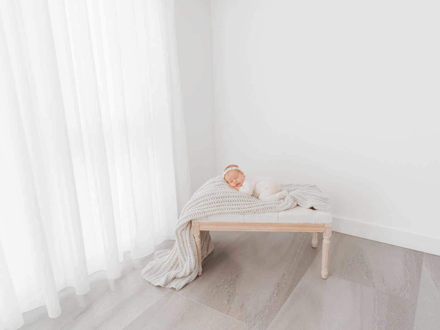 Newborn baby sleeping peacefully on a wooden bench draped with a soft gray blanket in a bright white studio.