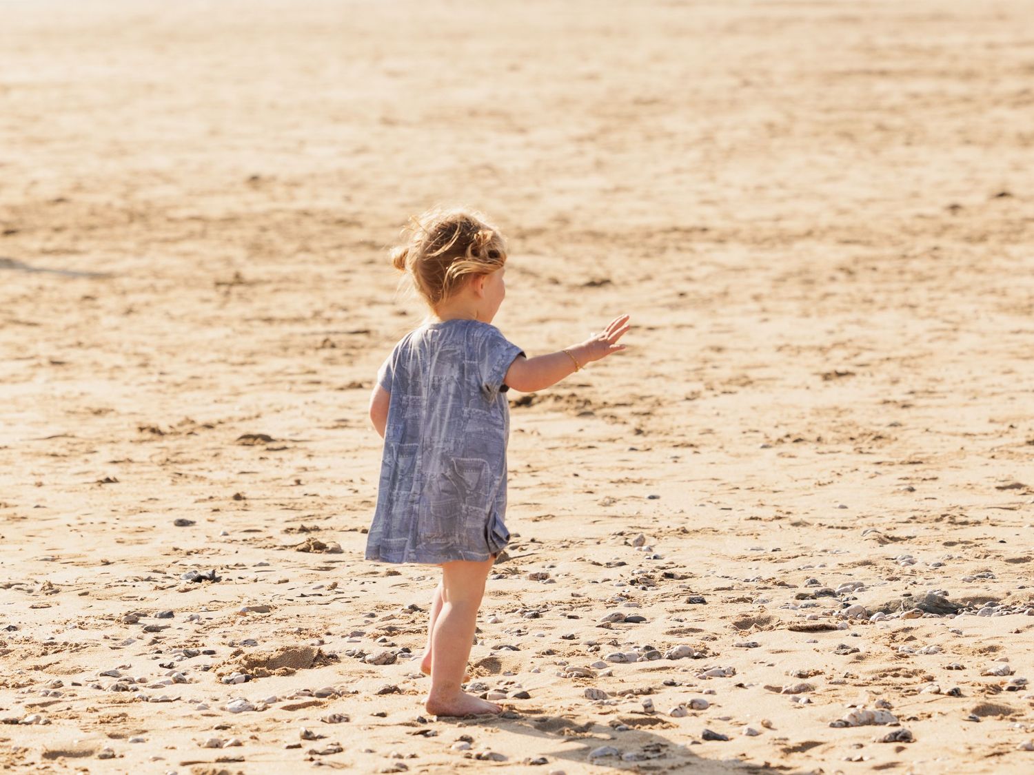 A small child in a blue dress stands barefoot on sandy ground while pointing into the distance at sunset.