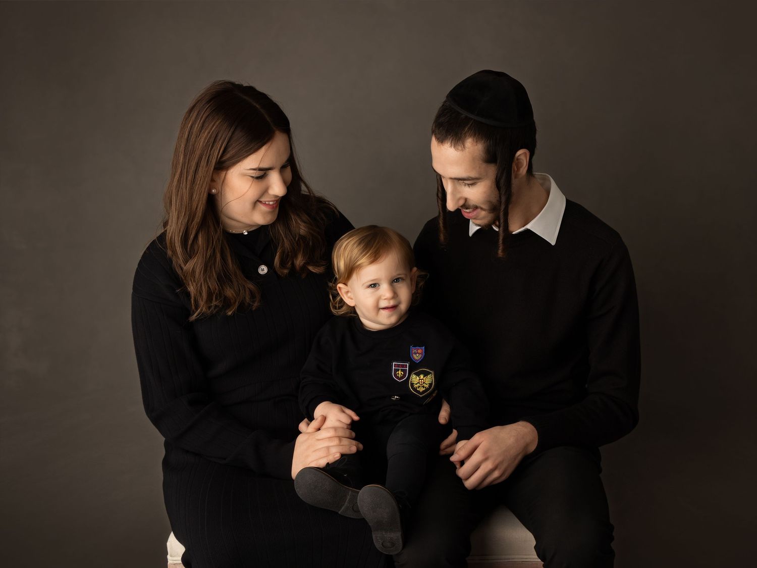 A loving family wearing matching black outfits poses together against a dark backdrop in a professional studio portrait.