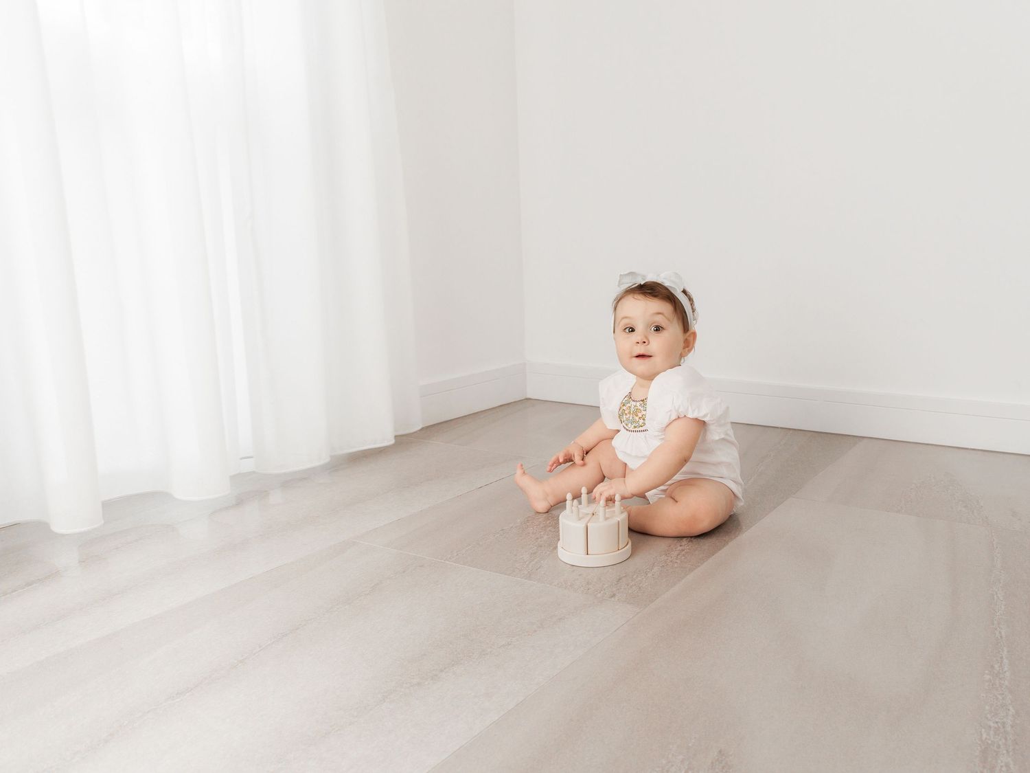 A baby in a white onesie sits on a light gray hardwood floor near sheer white curtains.