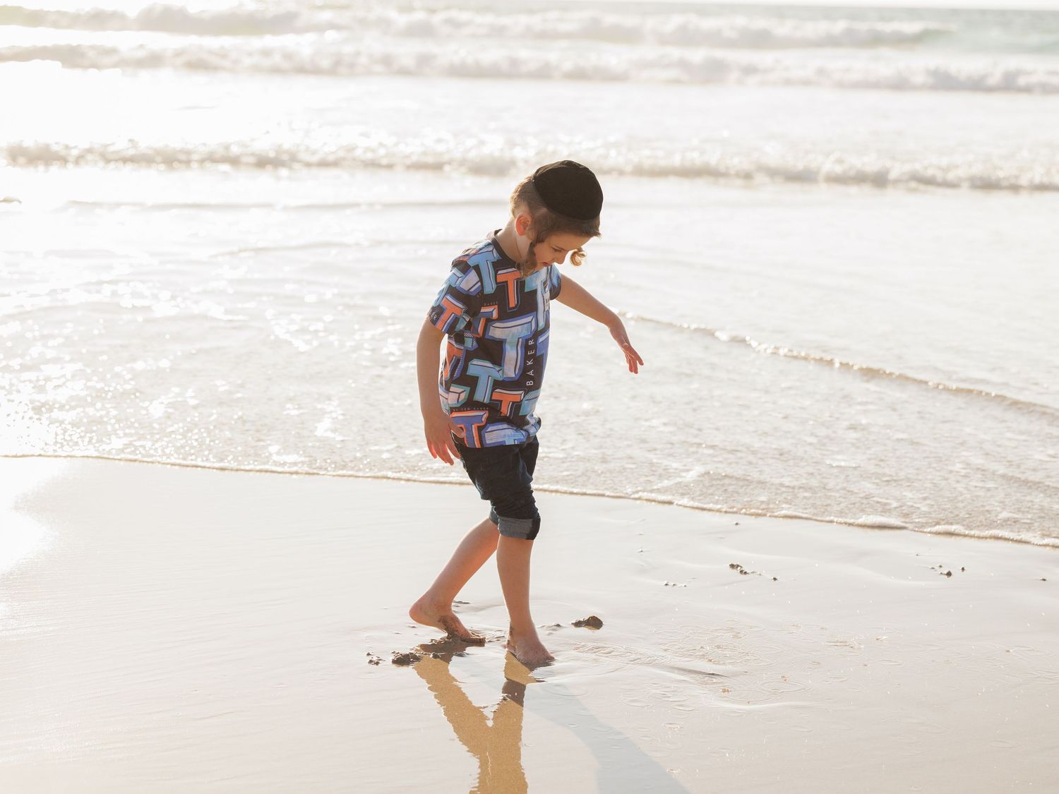 Barefoot child in plaid shirt walks along sandy beach shoreline with gentle waves in the background.