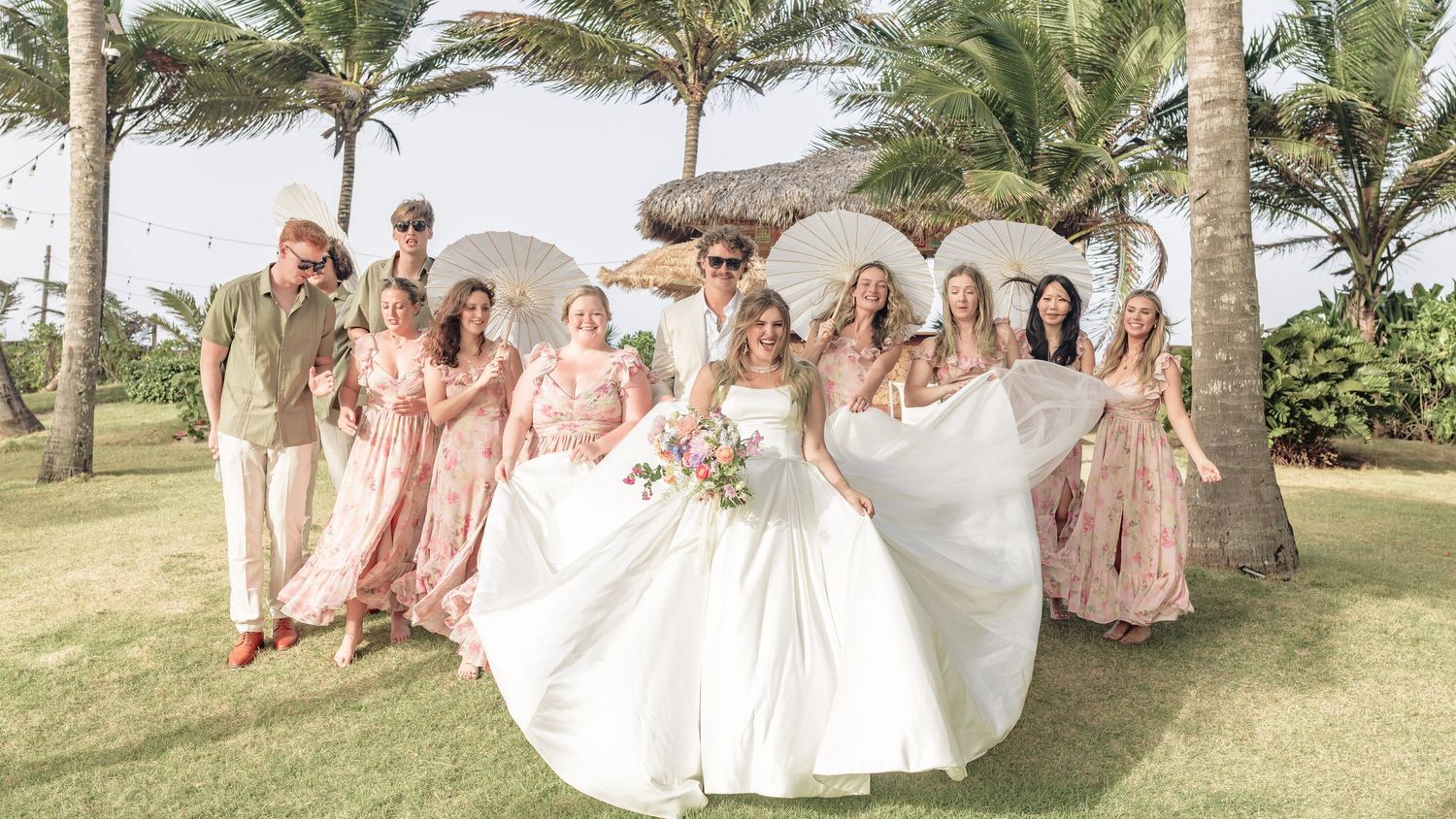 Wedding party poses together outdoors under palm trees in coordinated pink and white attire for group photos.