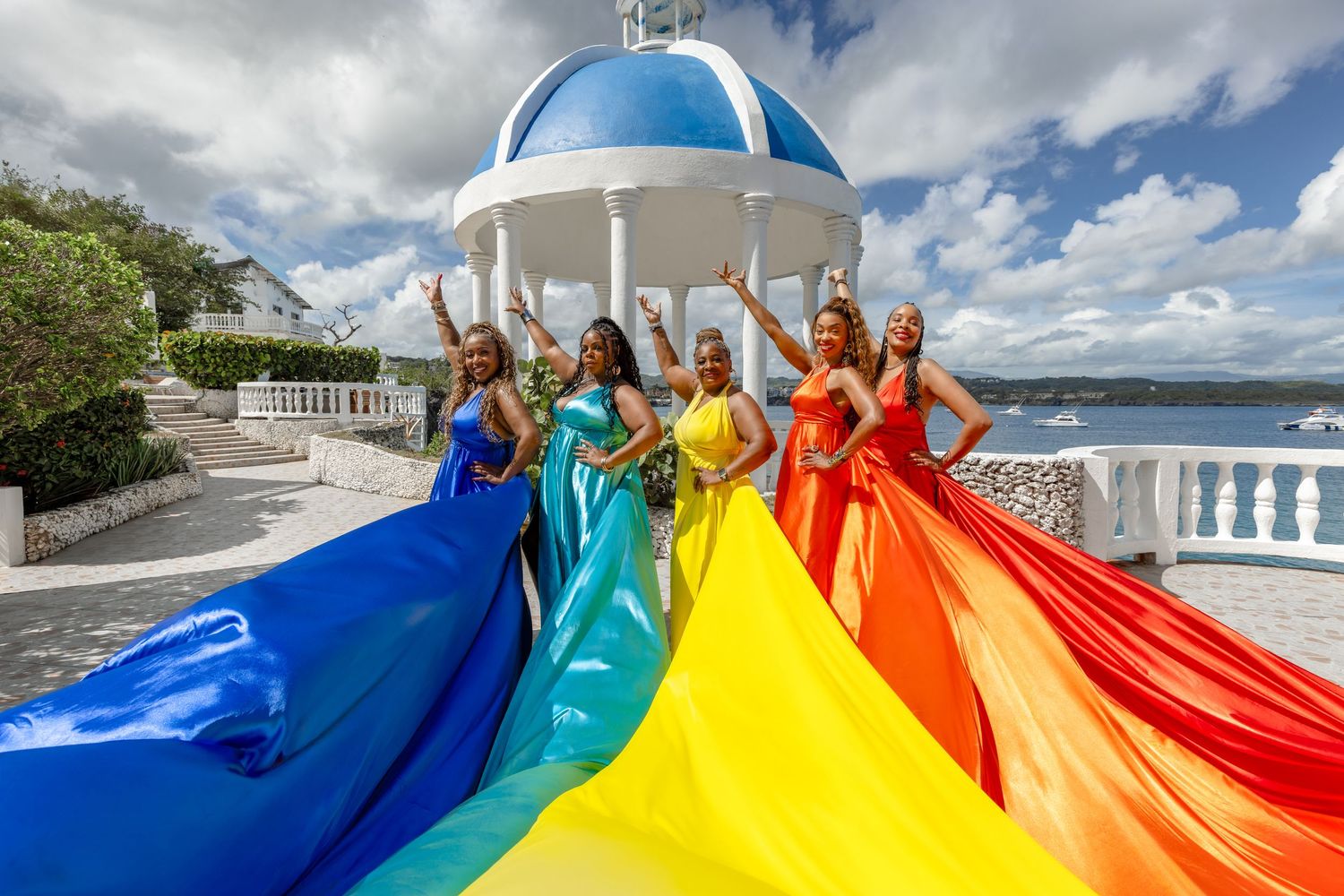 People hold a large rainbow flag while standing outdoors on a sunny day with blue skies and white clouds.