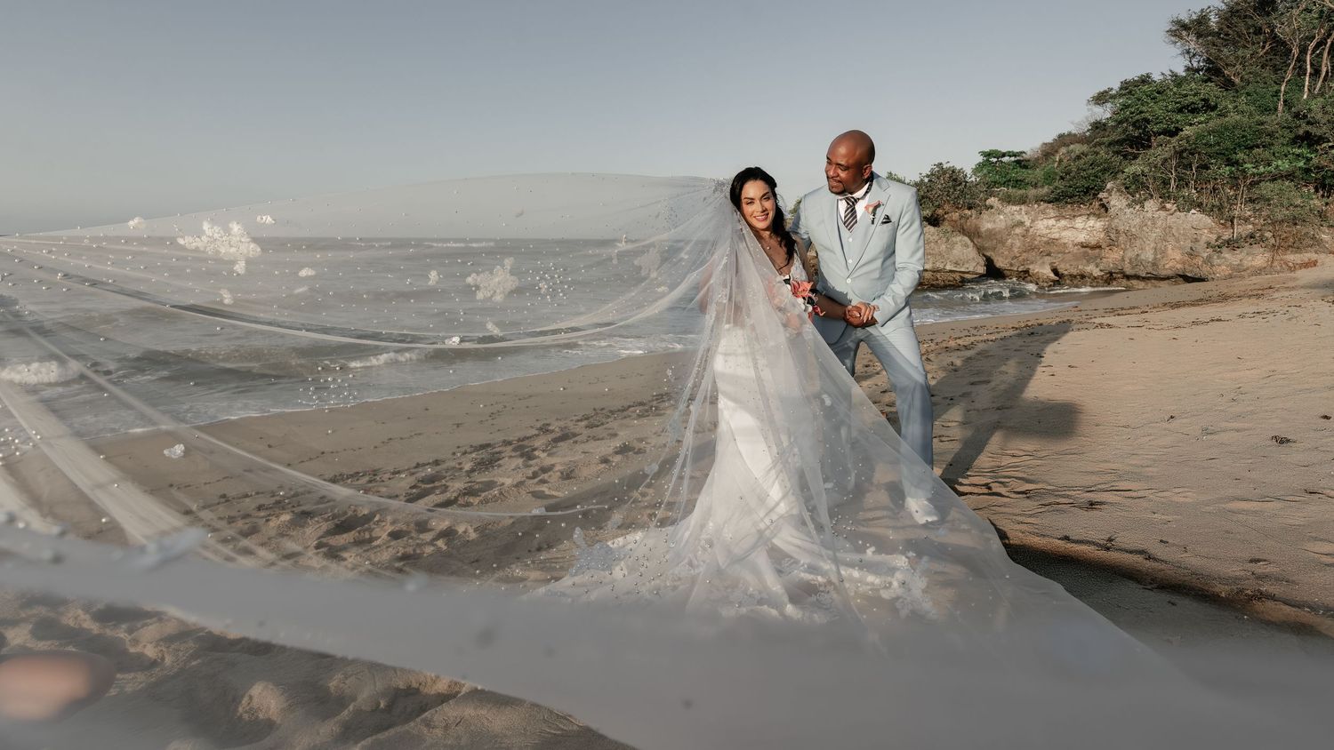 A couple in wedding attire walks along a misty beach shoreline at sunset during their romantic coastal photoshoot.