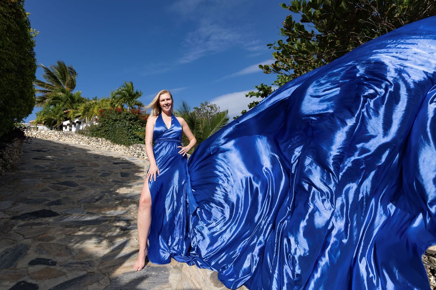 A flowing blue dress billows dramatically in the wind against a sunny outdoor backdrop with stone walls and greenery.