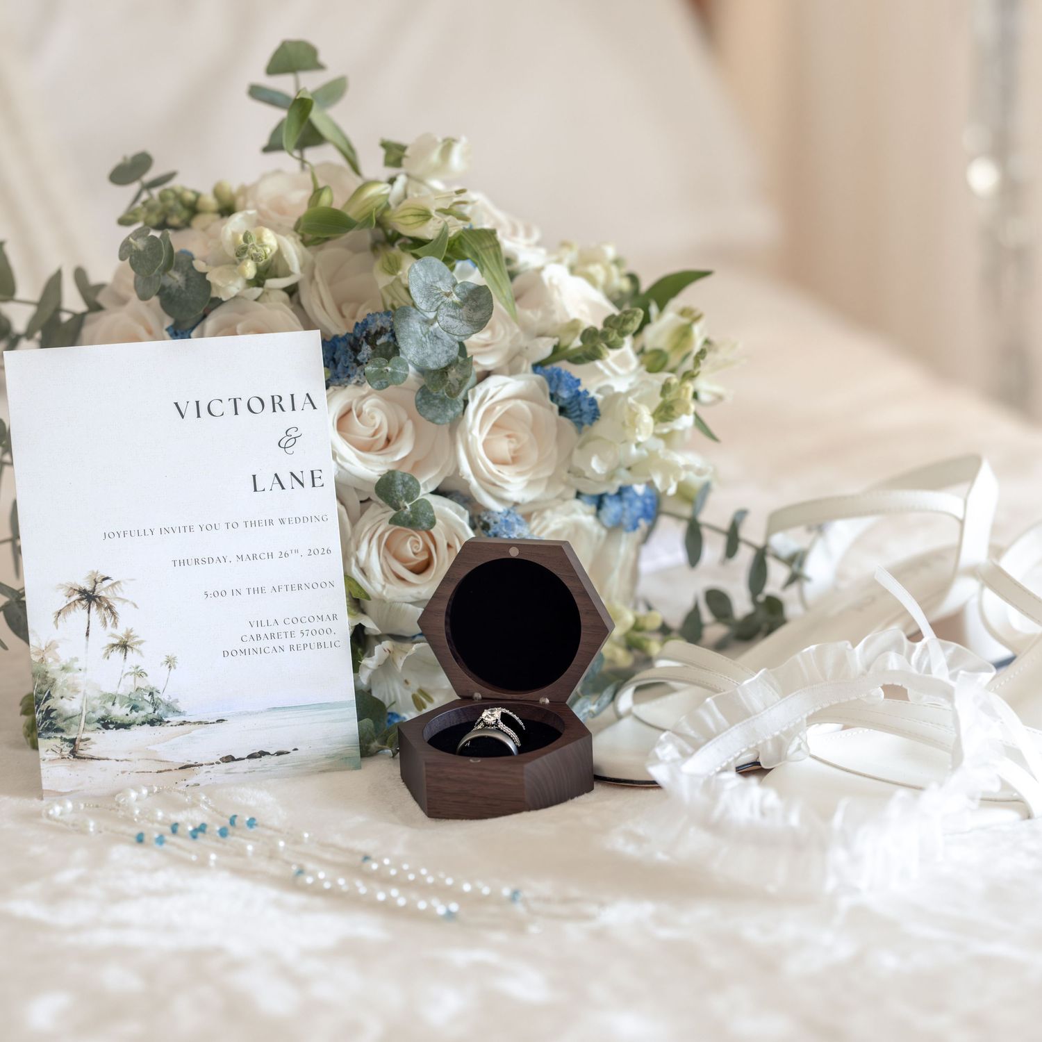 Wedding flat lay with floral bouquet, ring box, invitation cards, and ribbon on white fabric.