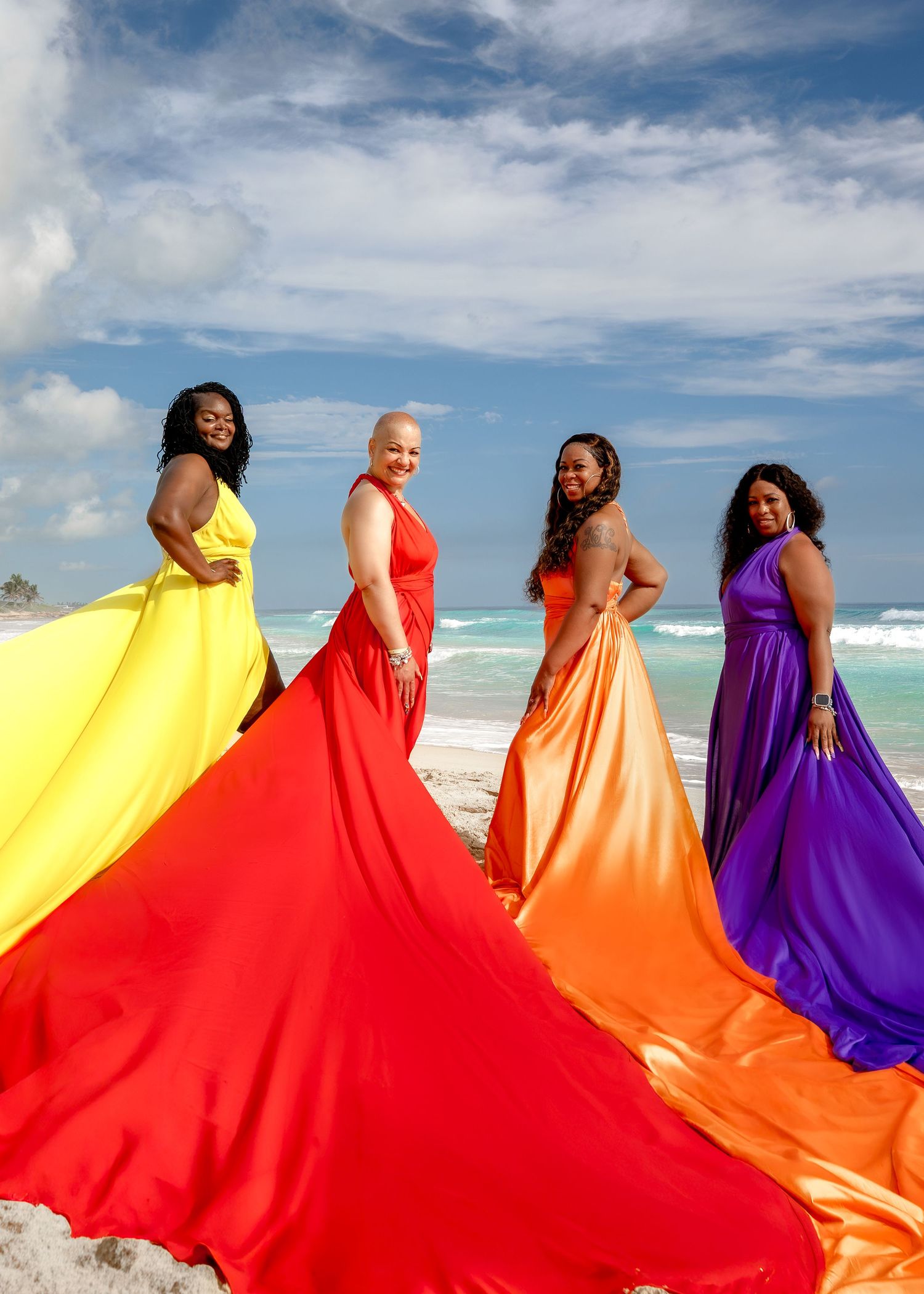 Four women in vibrant yellow, red, orange, and purple gowns pose on a sunny beach with flowing fabric.