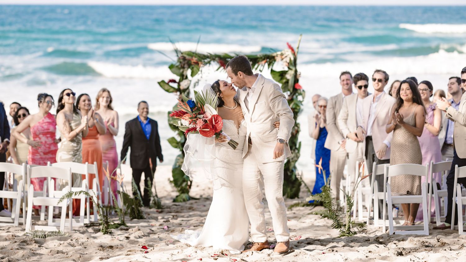 A wedding party gathers on a sunny beach with ocean waves and palm trees in the background.