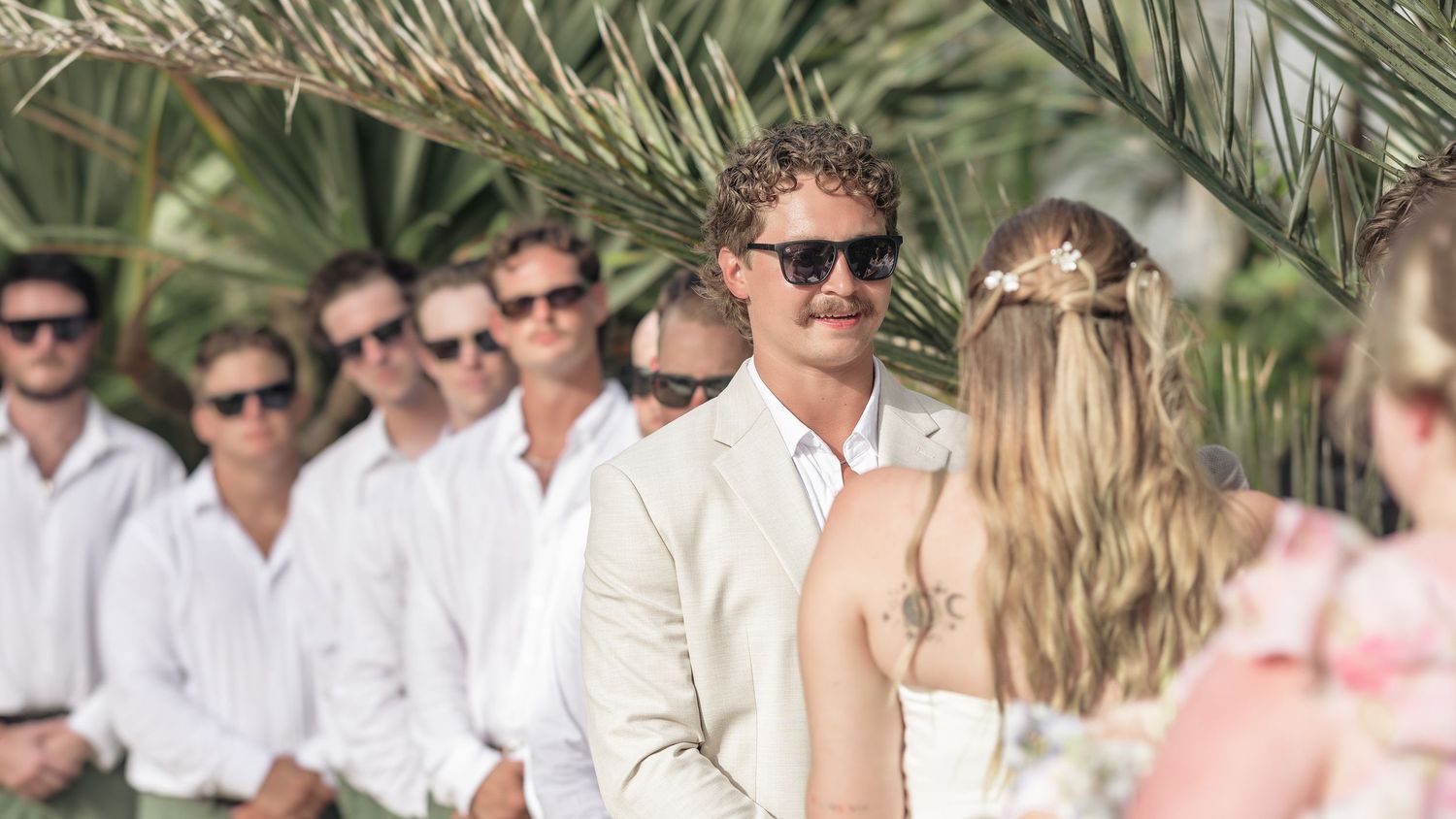 Wedding guests gather under palm trees at an outdoor tropical beach ceremony.