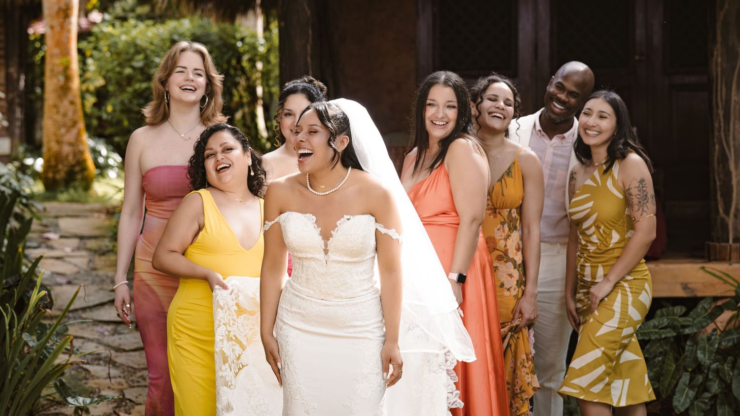 A wedding party in coordinated dresses walks together outdoors while laughing and smiling during a celebration.