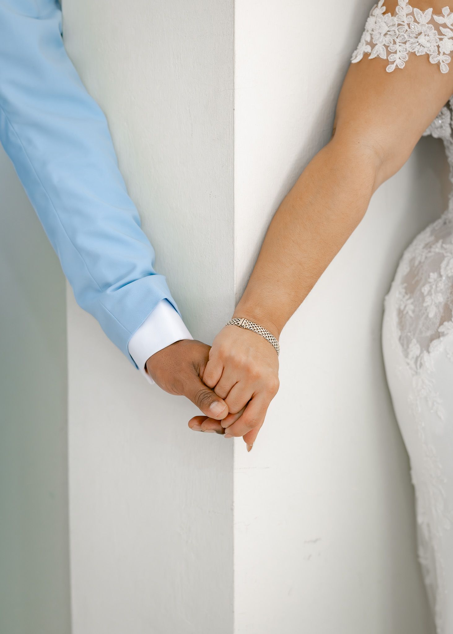 A couple in wedding attire holds hands against a white wall showing their rings and dress details.