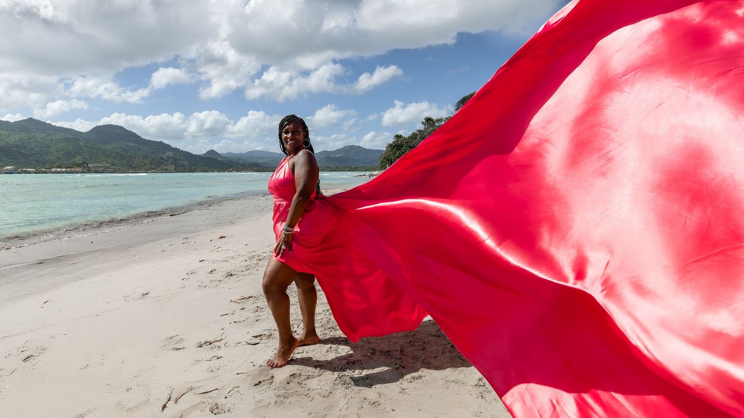 Red flowing fabric creates dramatic movement against a tropical beach landscape with turquoise water and blue sky.