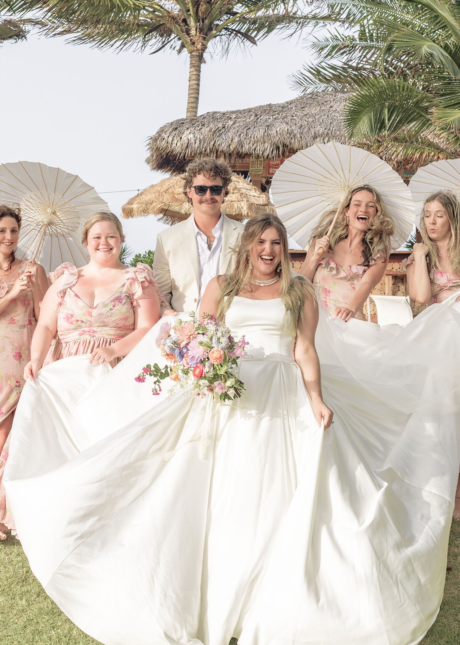 A group in white dresses poses with parasols under palm trees at a tropical beach wedding celebration.