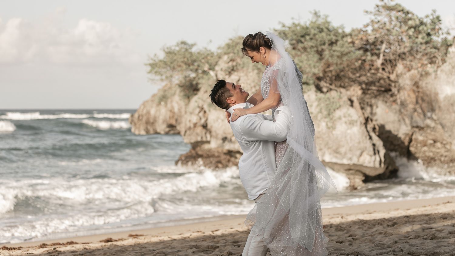 A couple embraces on a rocky beach during sunset with ocean waves crashing in the background.
