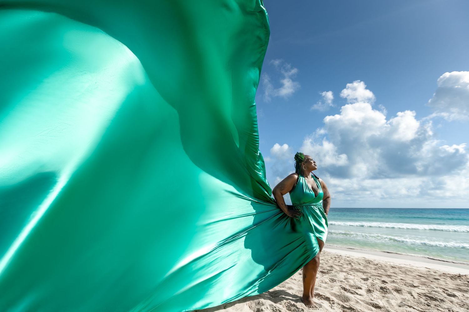 Person in turquoise dress stands on beach holding flowing fabric against blue sky and ocean backdrop.