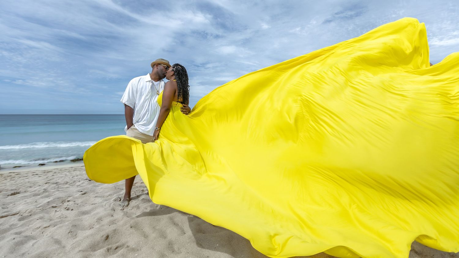 A bright yellow fabric billows gracefully in the wind along a sandy beach against a blue sky and ocean backdrop.
