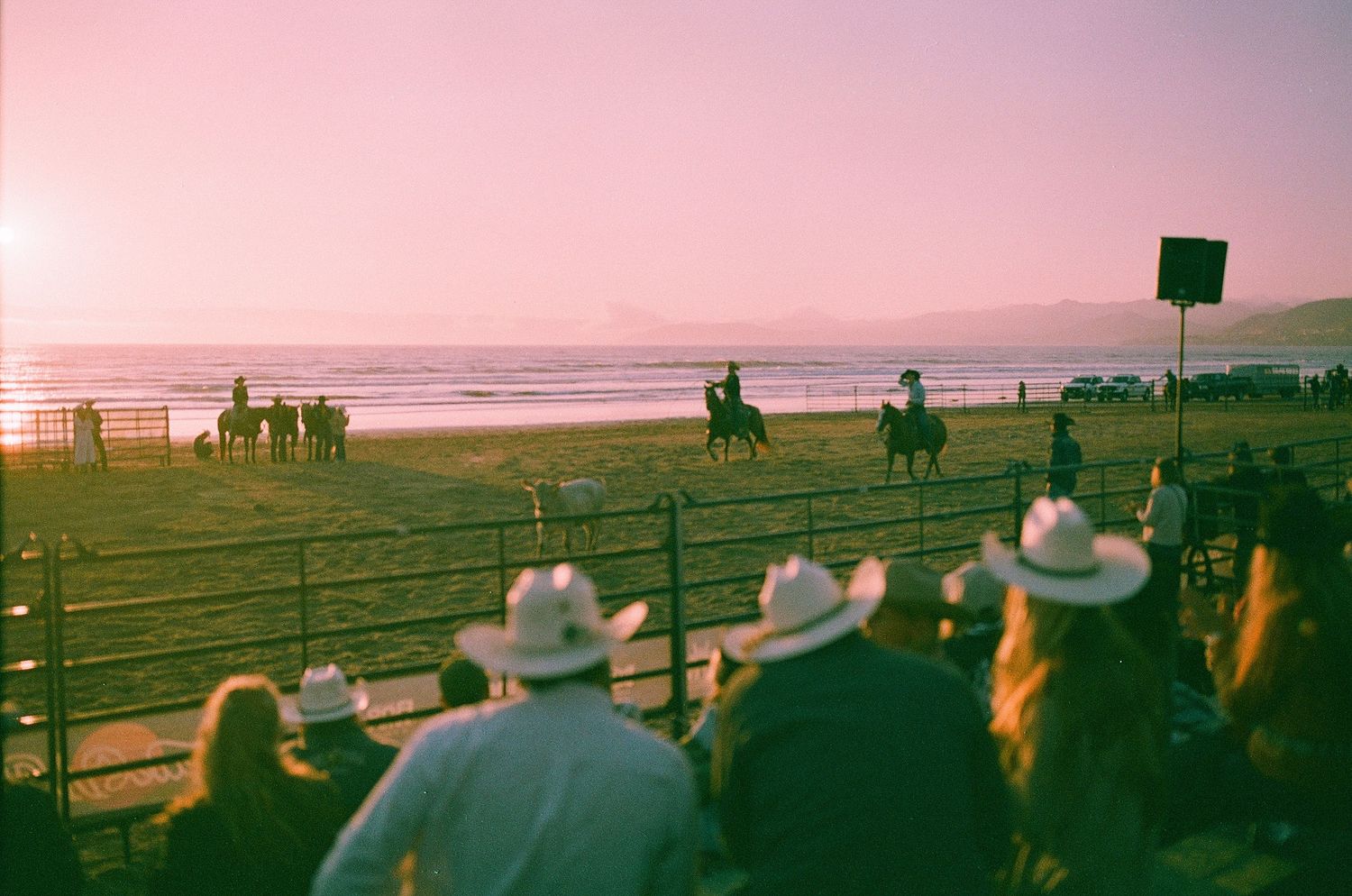 Cowboys on horseback ride along a beach fence at sunset while spectators in cowboy hats watch the coastal rodeo event.