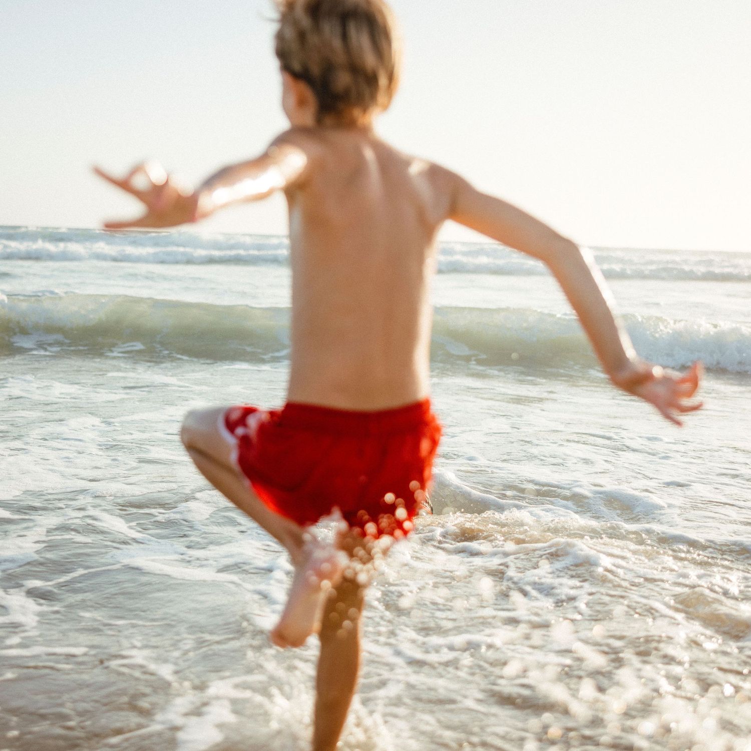 Young boy in red swim trunks running joyfully into ocean waves on a sunny beach day.