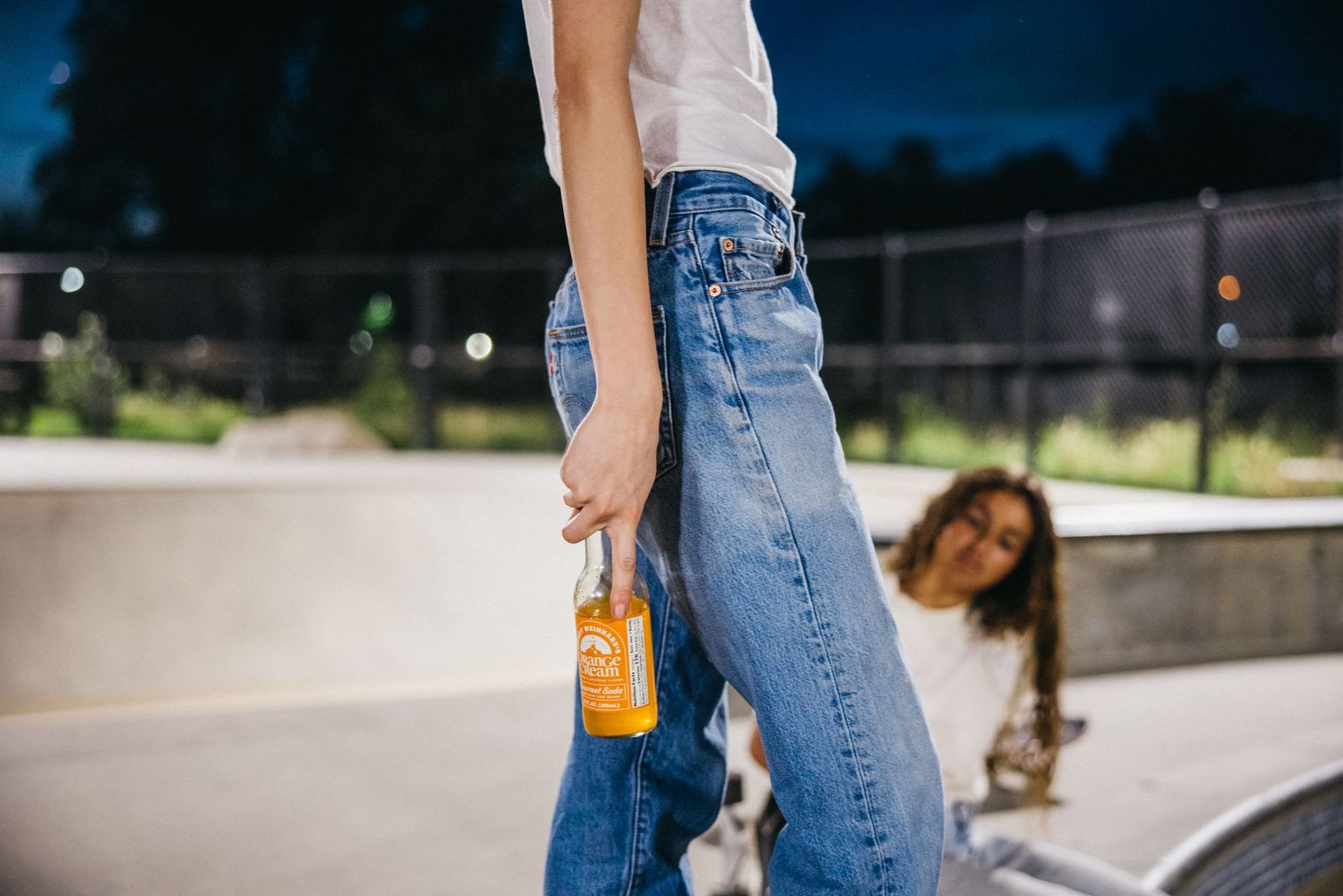Young woman in high-waisted jeans holding an orange drink at a skate park.