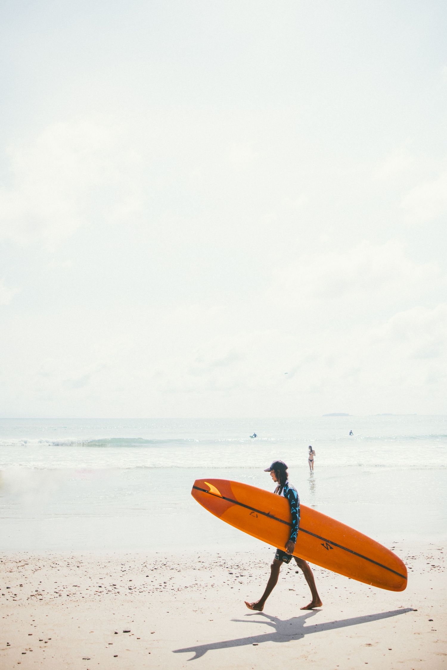 A surfer carries an orange longboard along a sandy beach with gentle waves and a bright cloudy sky.