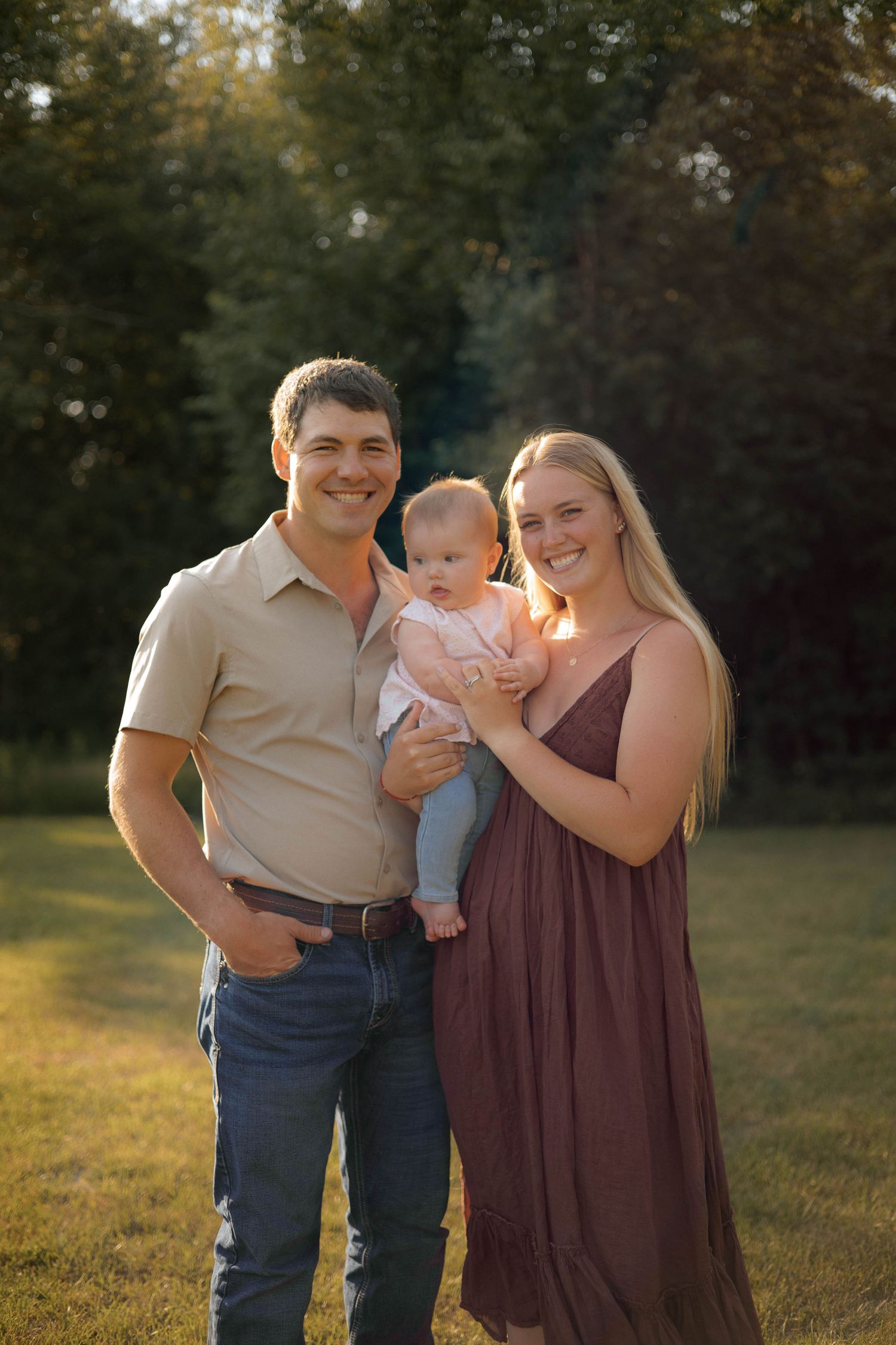 Young family portrait outdoors at golden hour, parents smiling holding infant baby in pink, green trees background.