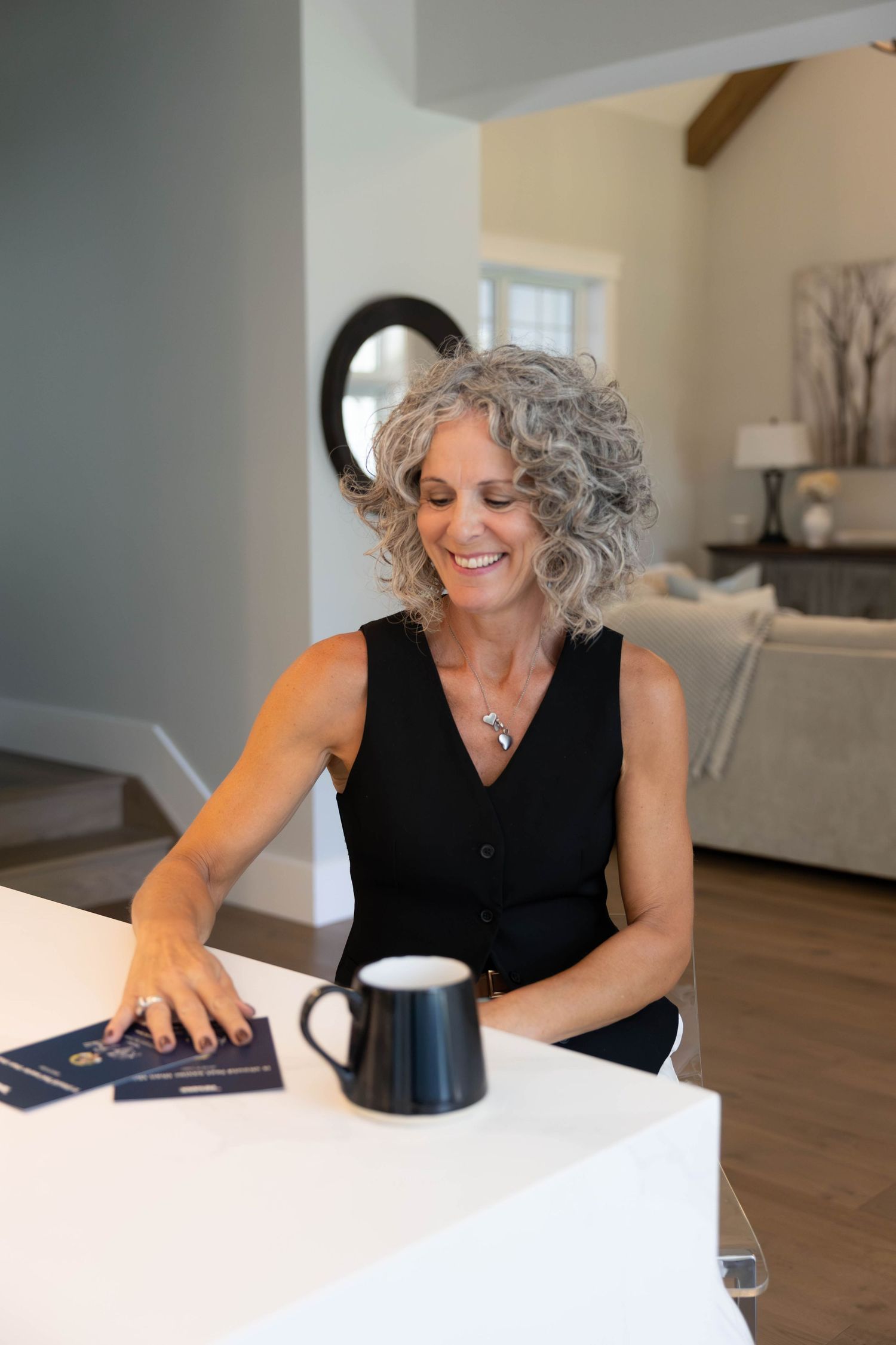 Smiling woman with curly gray hair sits at white counter with coffee mug and documents in modern home interior.