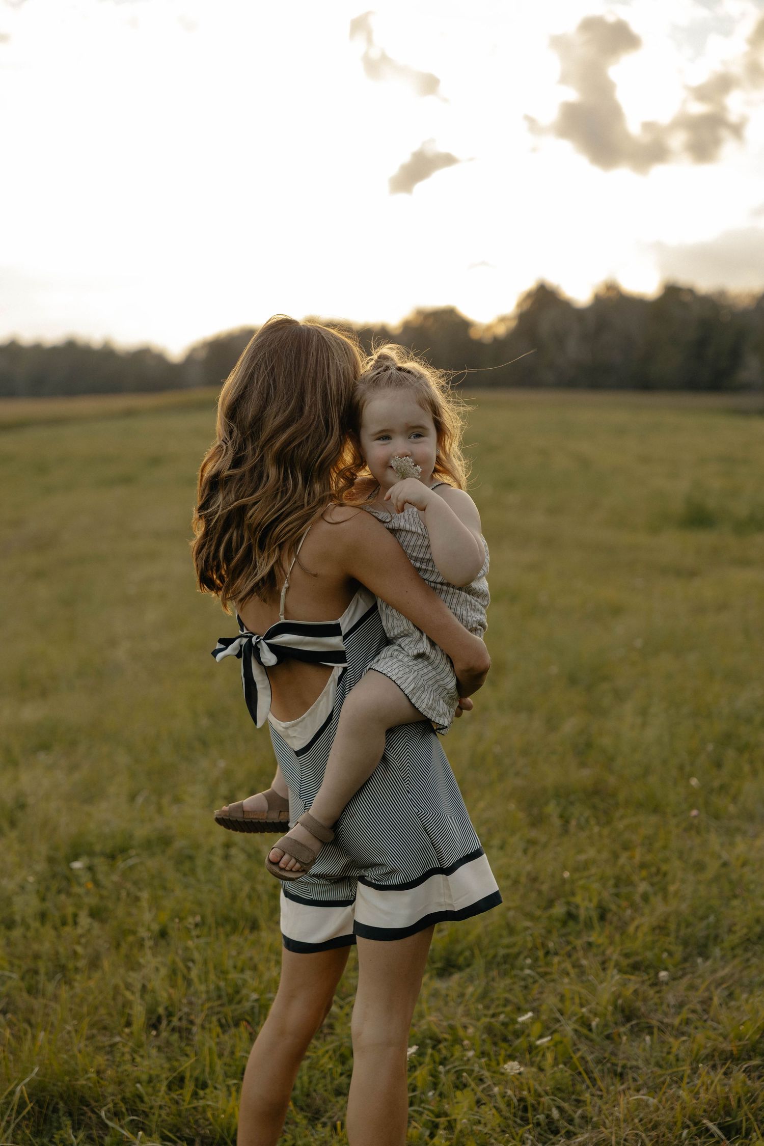 A mother holds her toddler in a sunlit green field at golden hour, both wearing matching outfits.