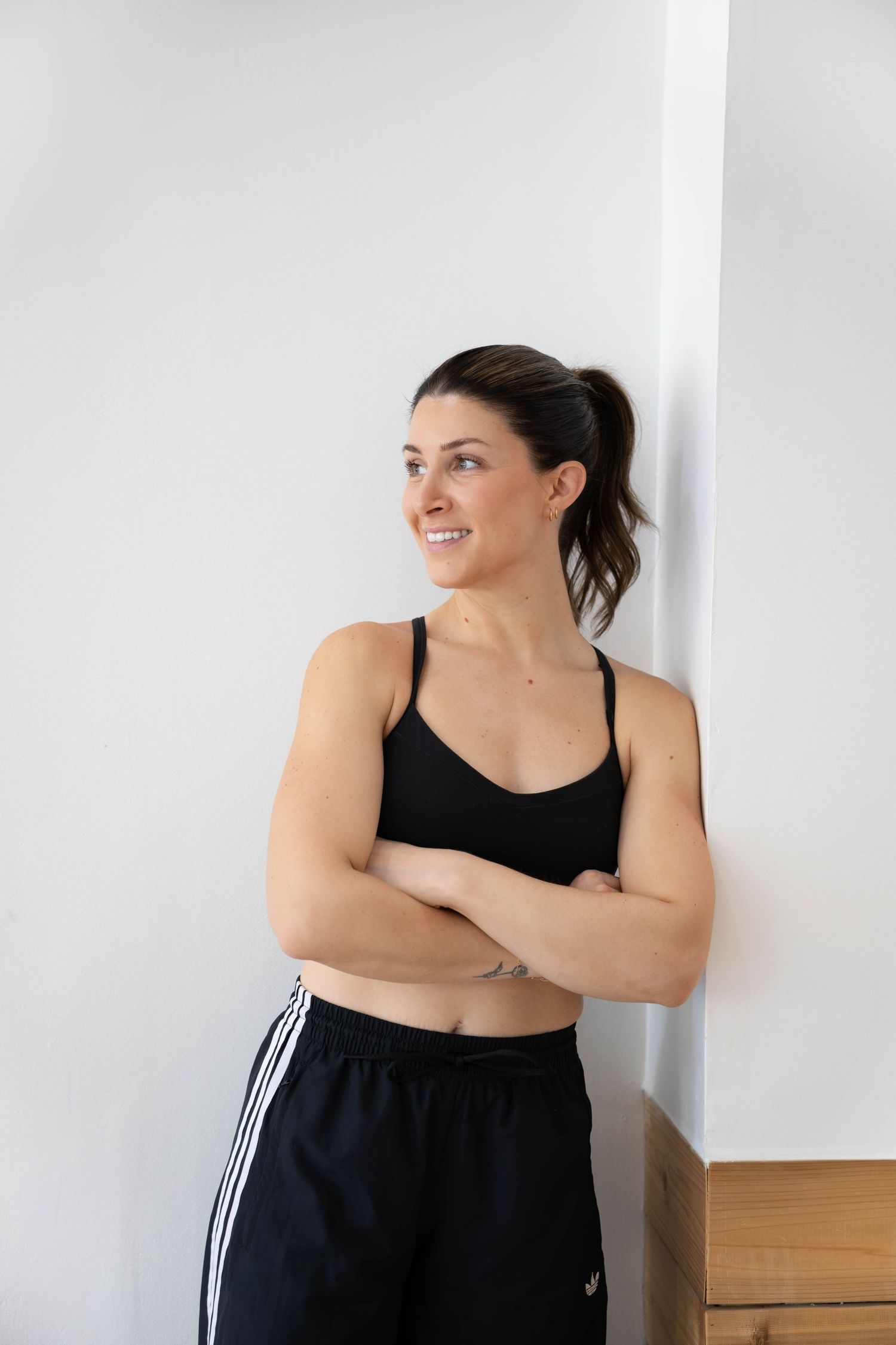 Woman in black sports bra and Adidas track pants leaning against white wall with arms crossed, smiling.