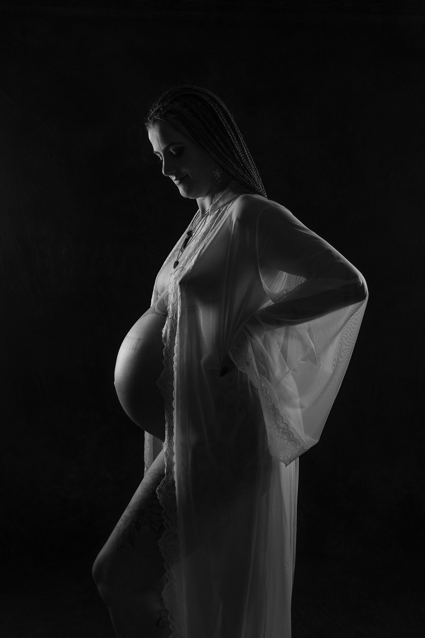 Black and white maternity photo of pregnant woman in sheer white robe posing against dark background.