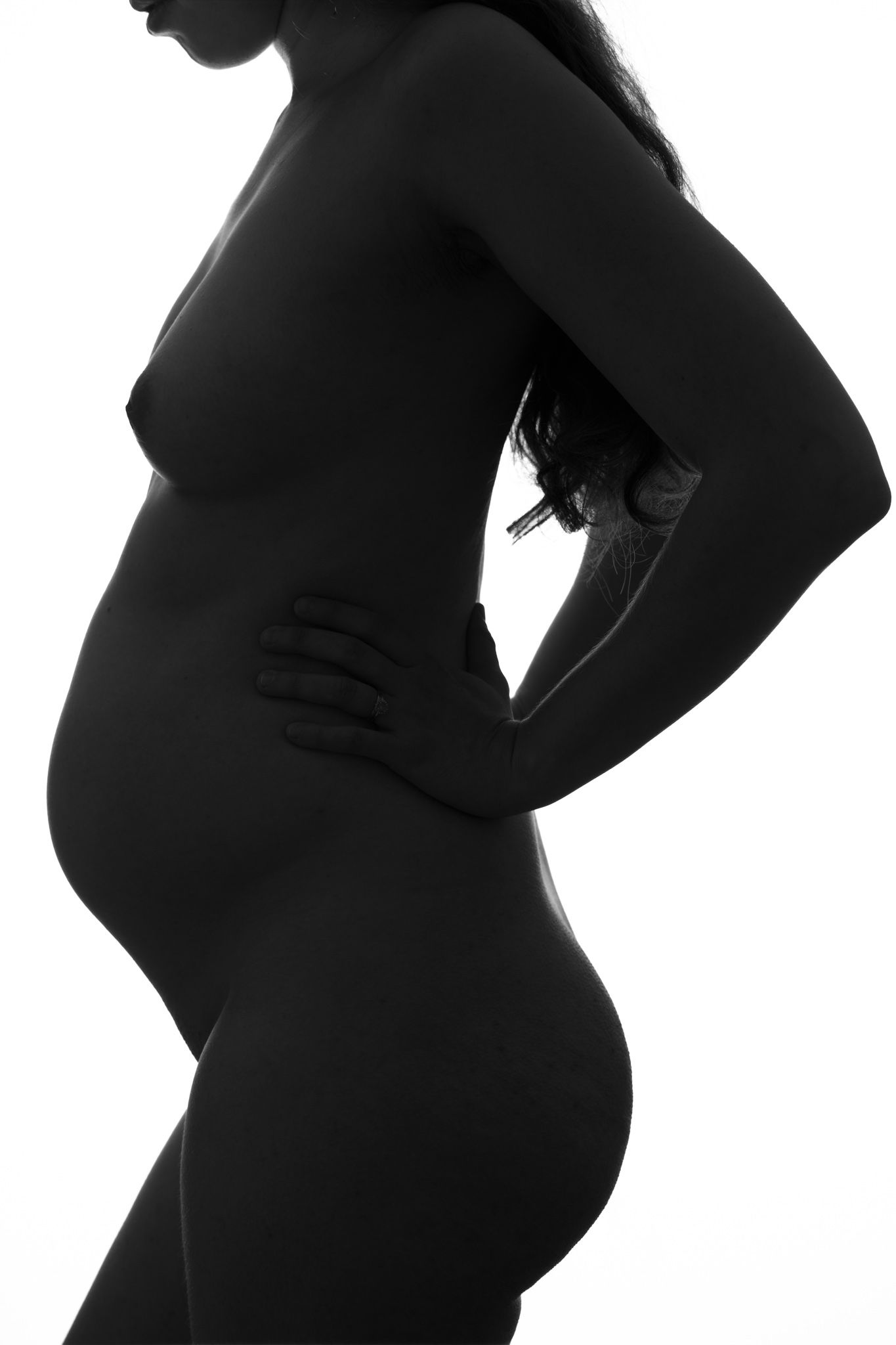 Black and white silhouette of a pregnant woman standing sideways, cradling her baby bump against white background. Boudoir photography