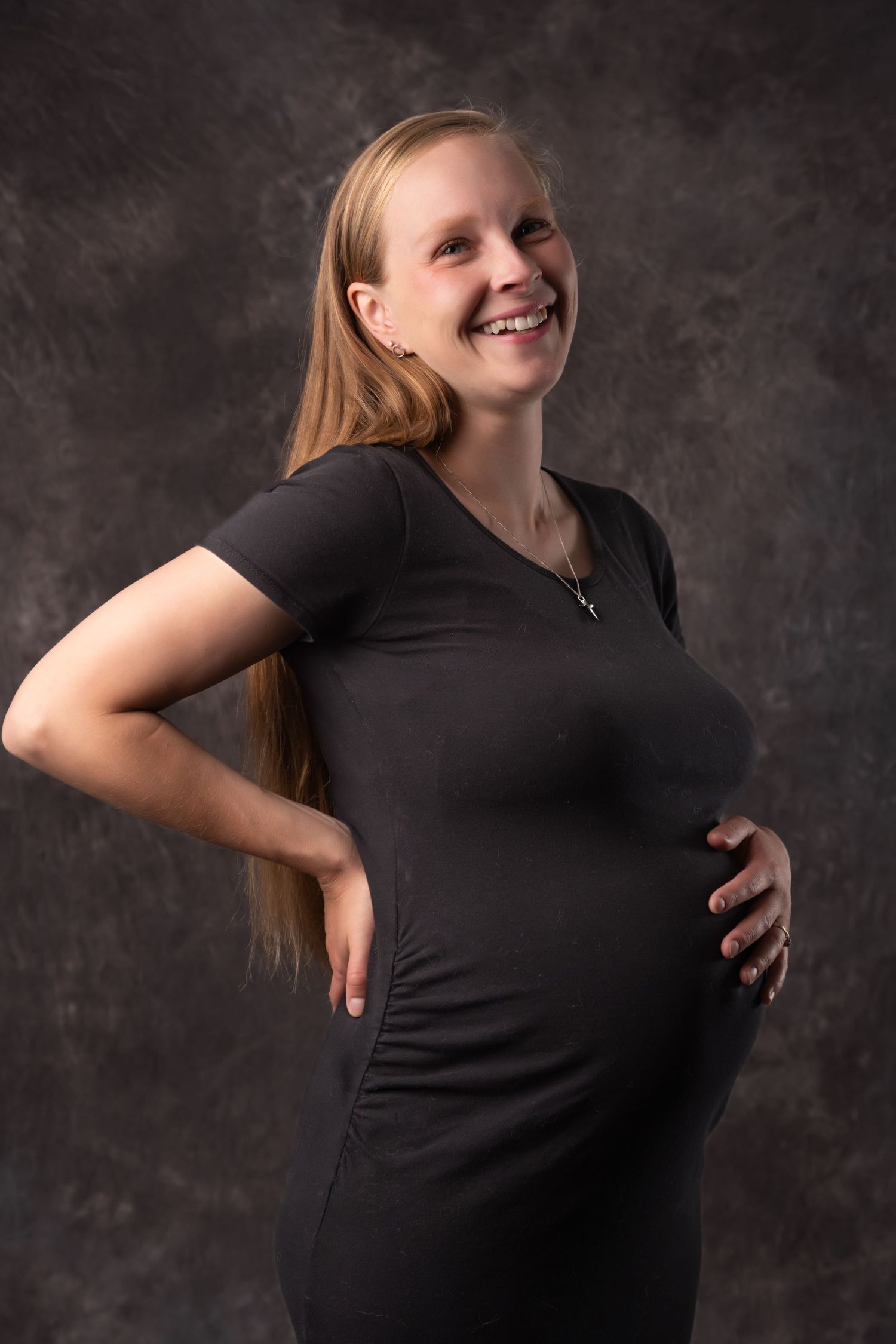Happy pregnant blonde woman in black dress smiles holding baby bump in studio maternity photo. Boudoir photography Christchurch maternity
