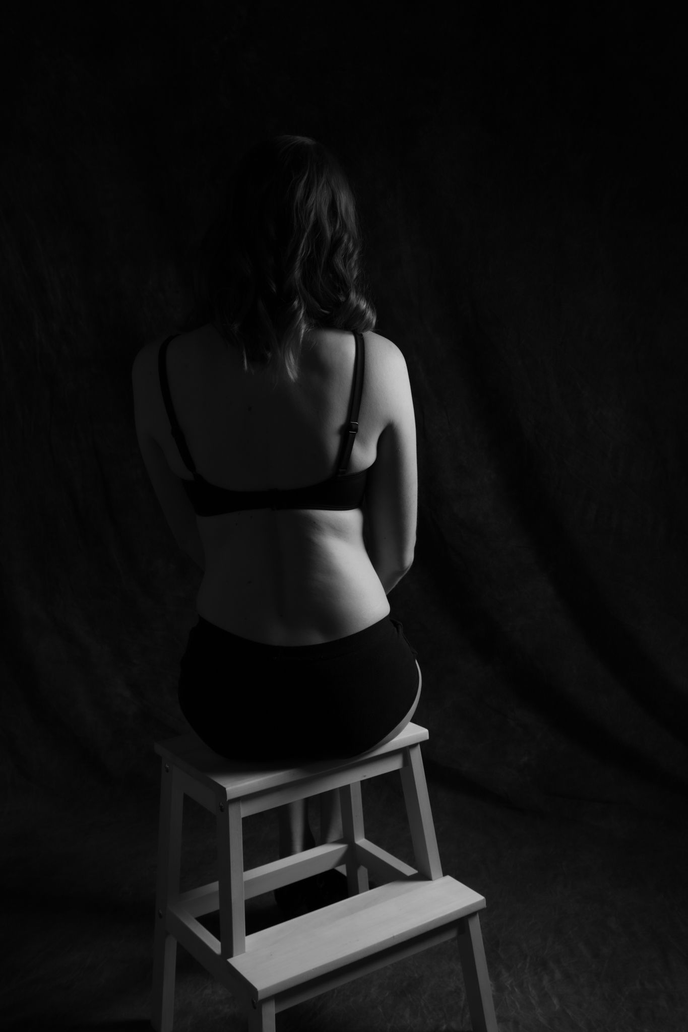 Black and white rear view of a woman in dark bra and underwear sitting on a white stool against a dark backdrop. Boudoir photography Christc