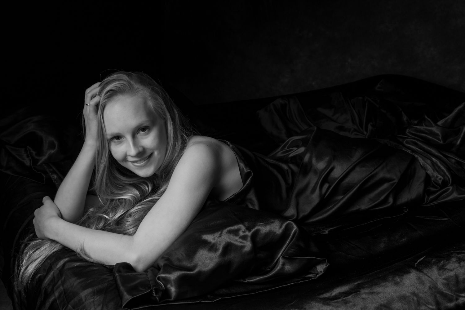 Black and white portrait of a smiling blonde woman lying on dark satin sheets with arms crossed. Boudoir photography Christchurch