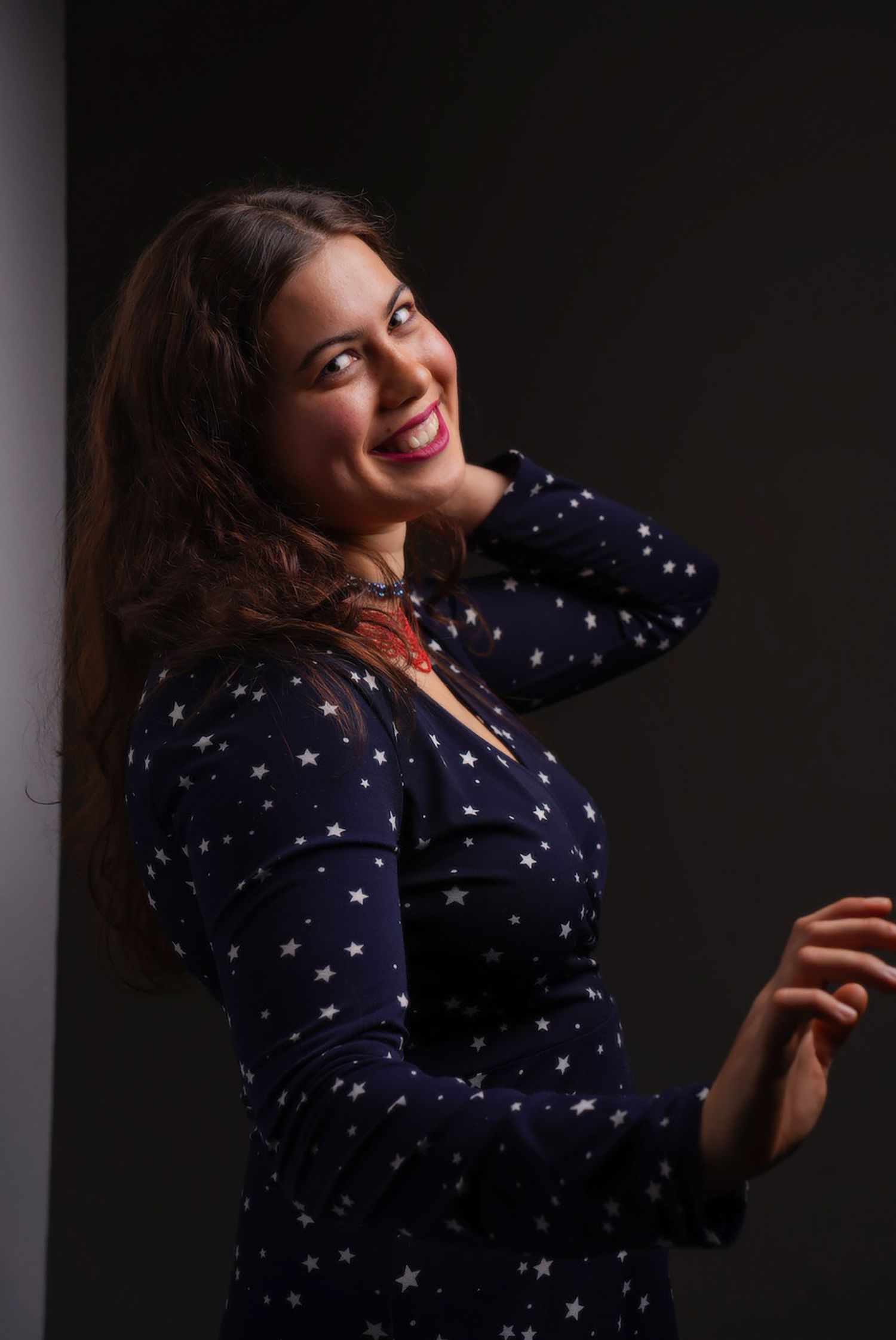 Smiling woman in navy star print dress posing against dark background with hand behind head. portrait photography Christchurch