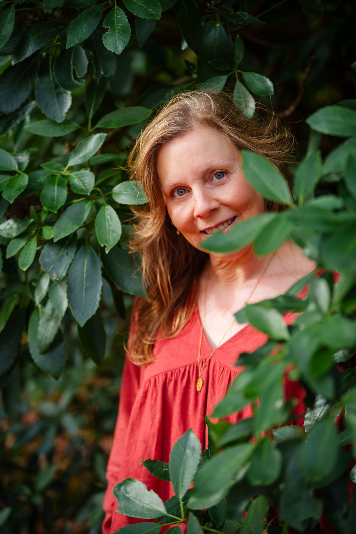 Smiling woman with blue eyes peering through lush green leaves wearing a red dress outdoors. portrait photography Christchurch