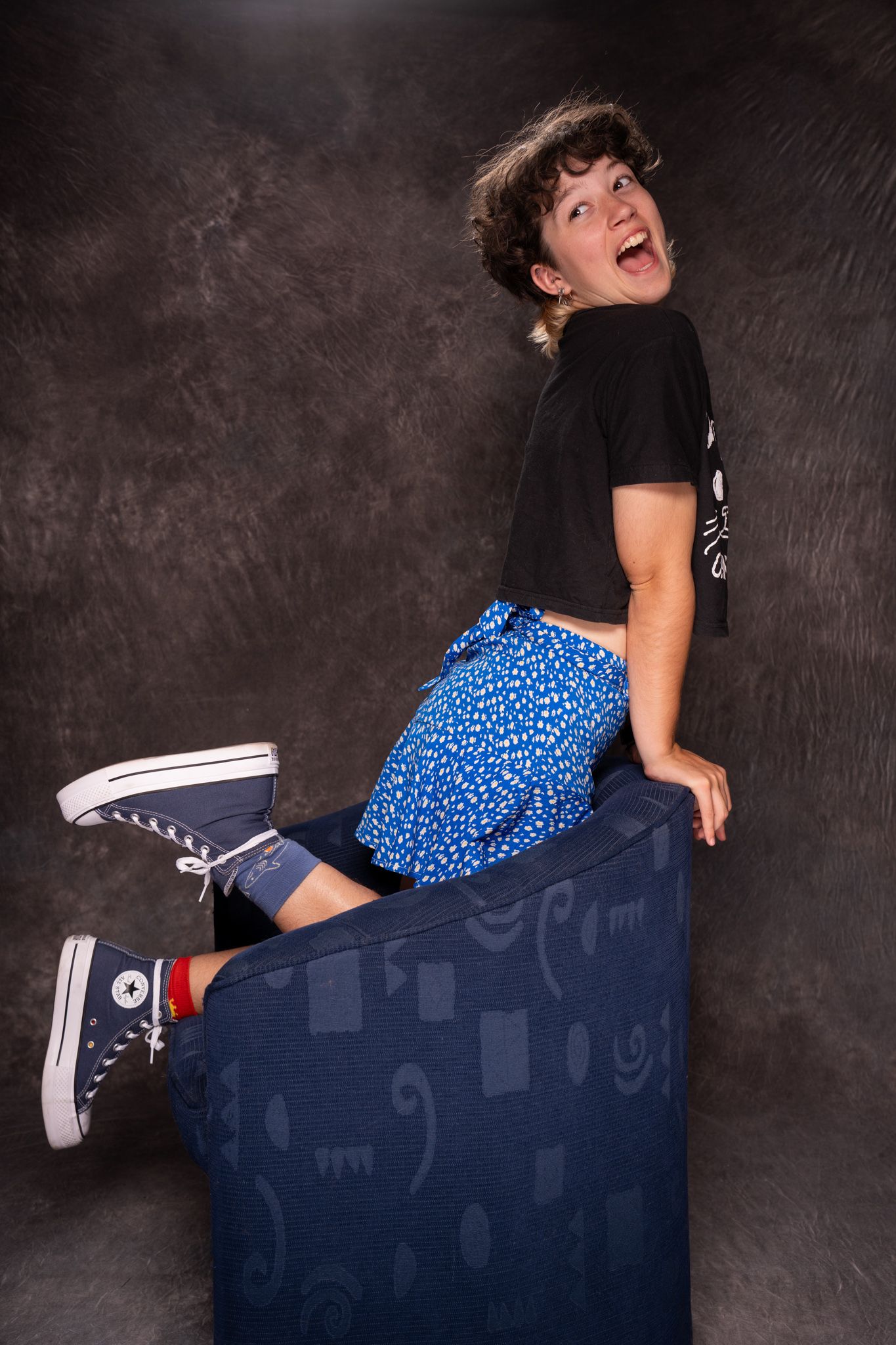 Energetic person in black tee, blue shorts and Converse sneakers climbing on a chair with an excited expression. portrai photography Christc