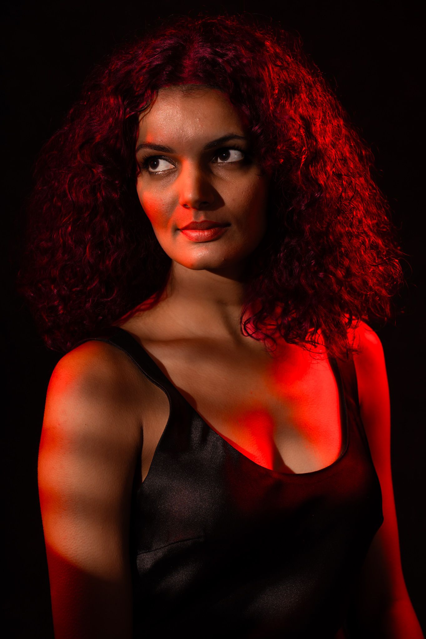 Woman with curly red hair in black dress illuminated by dramatic red lighting against dark background. Boudoir photography Christchurch