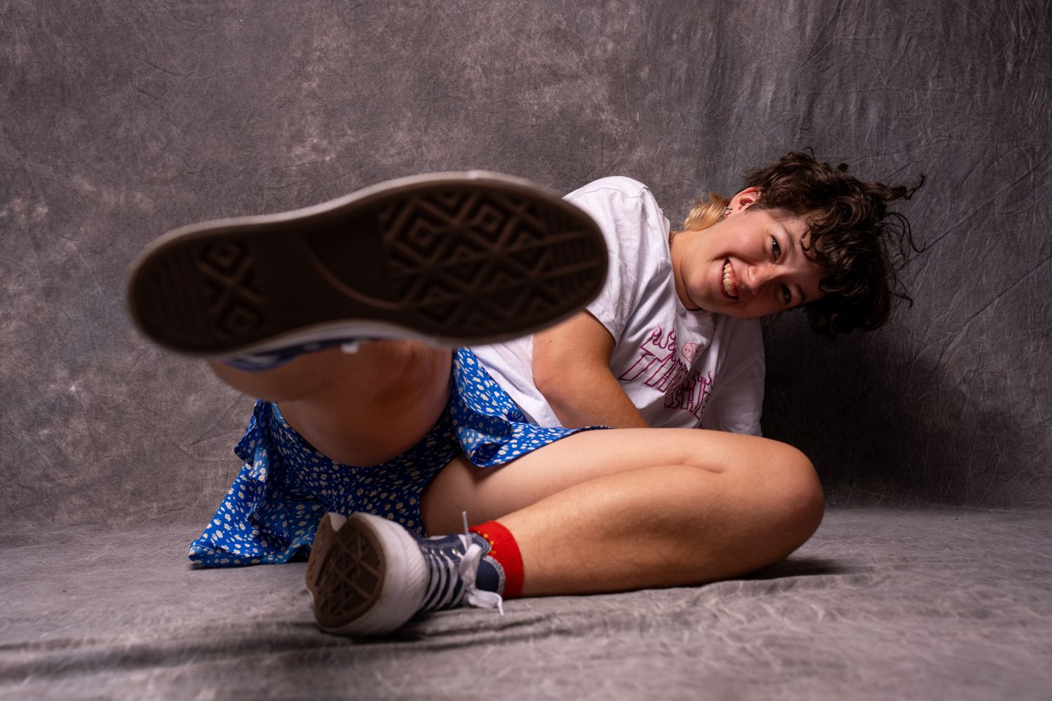 Playful young woman sitting on floor in blue sequin skirt and white tee, kicking up Converse sneaker. portrait photography Christchurch