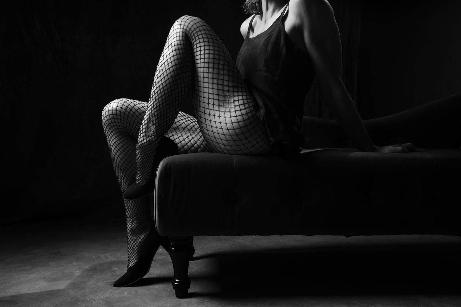 Black and white artistic photo of a woman in fishnet stockings and heels seated on a dark sofa. Boudoir photography Christchurch