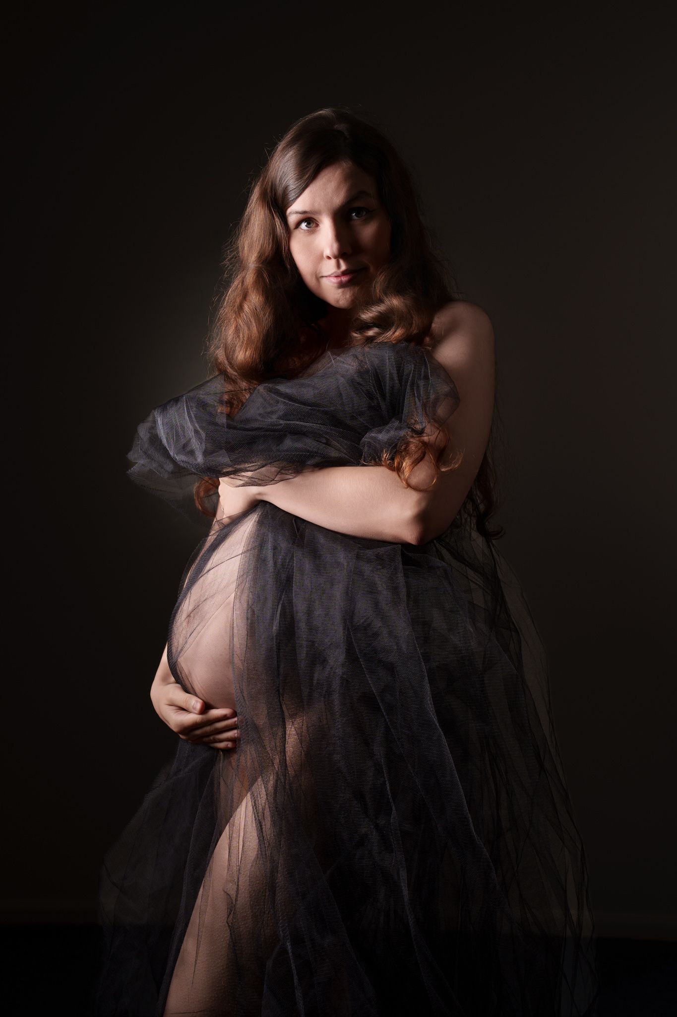 Pregnant woman wrapped in sheer dark fabric posing in moody studio lighting.  Boudoir photography Christchurch maternity