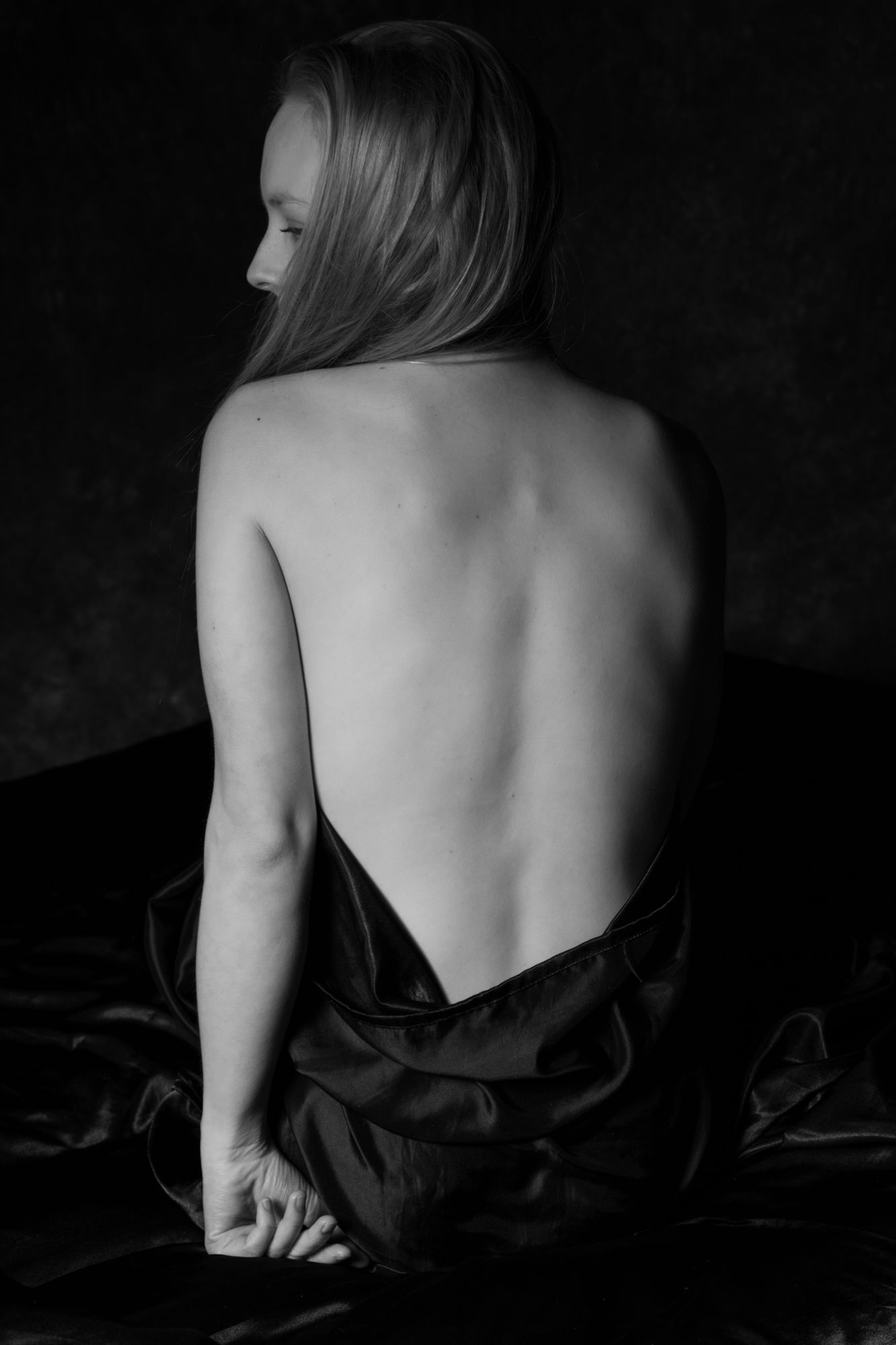 Black and white artistic portrait of a woman with bare back sitting on dark satin fabric. Boudoir photography Christchurch