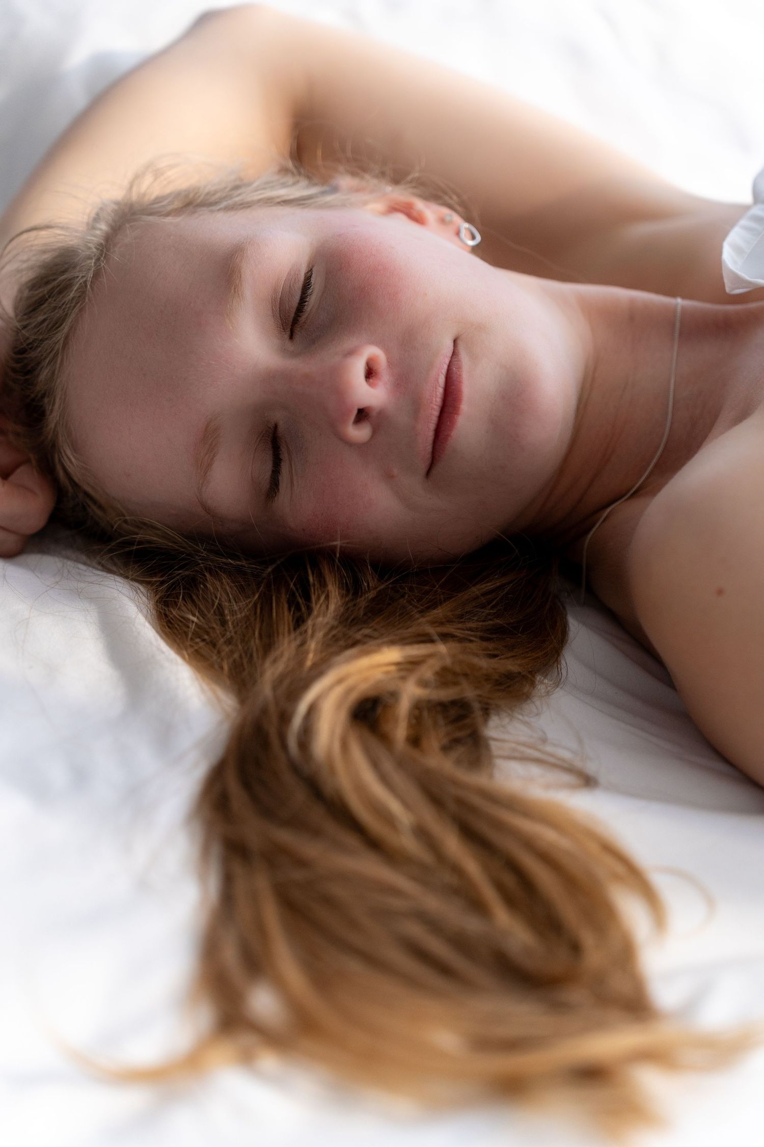 Young woman with long hair lying on white bedding with eyes closed, appearing peaceful and relaxed.