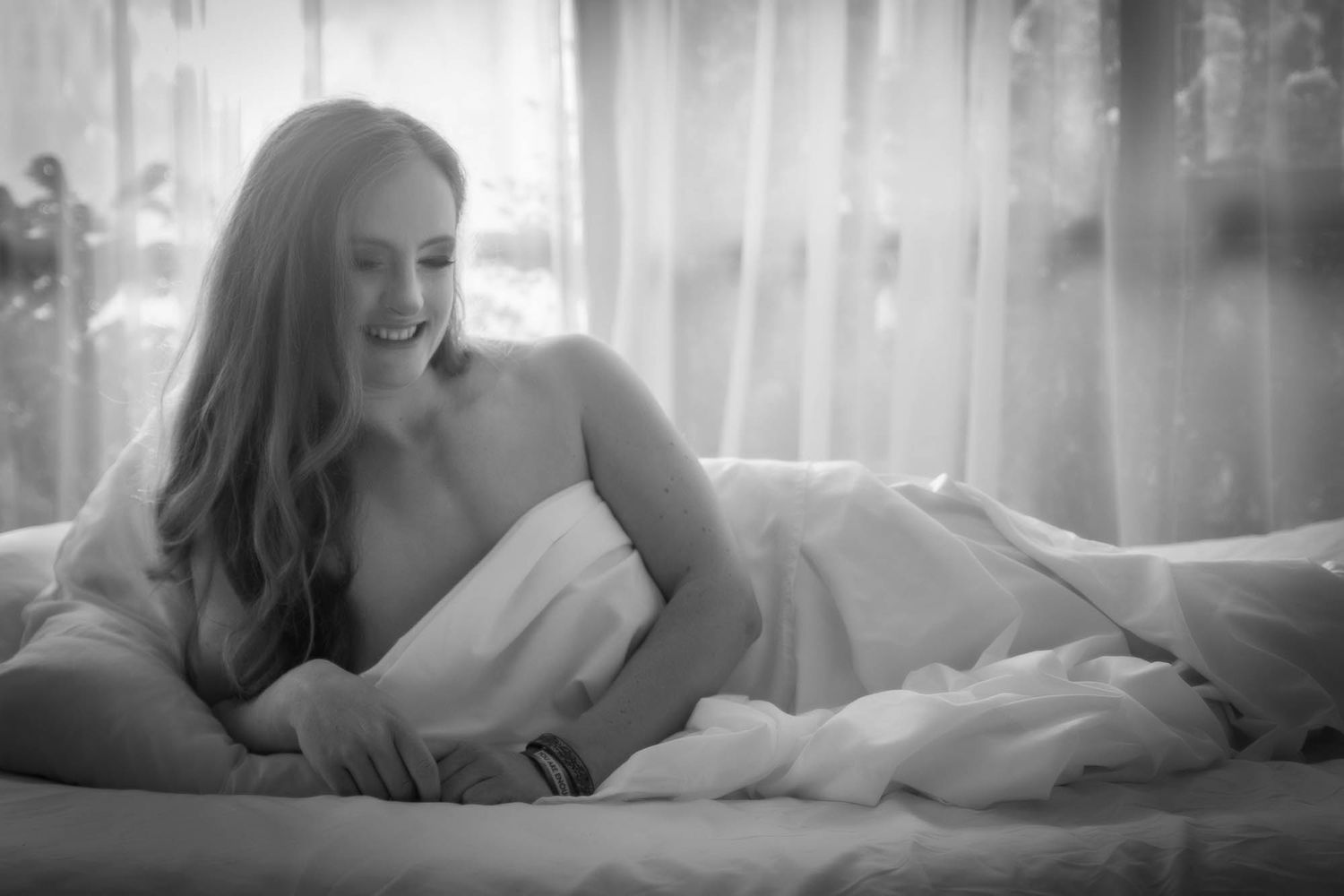 Black and white boudoir photo of smiling woman with long hair lying on bed by bright window. Boudoir photography Christchurch