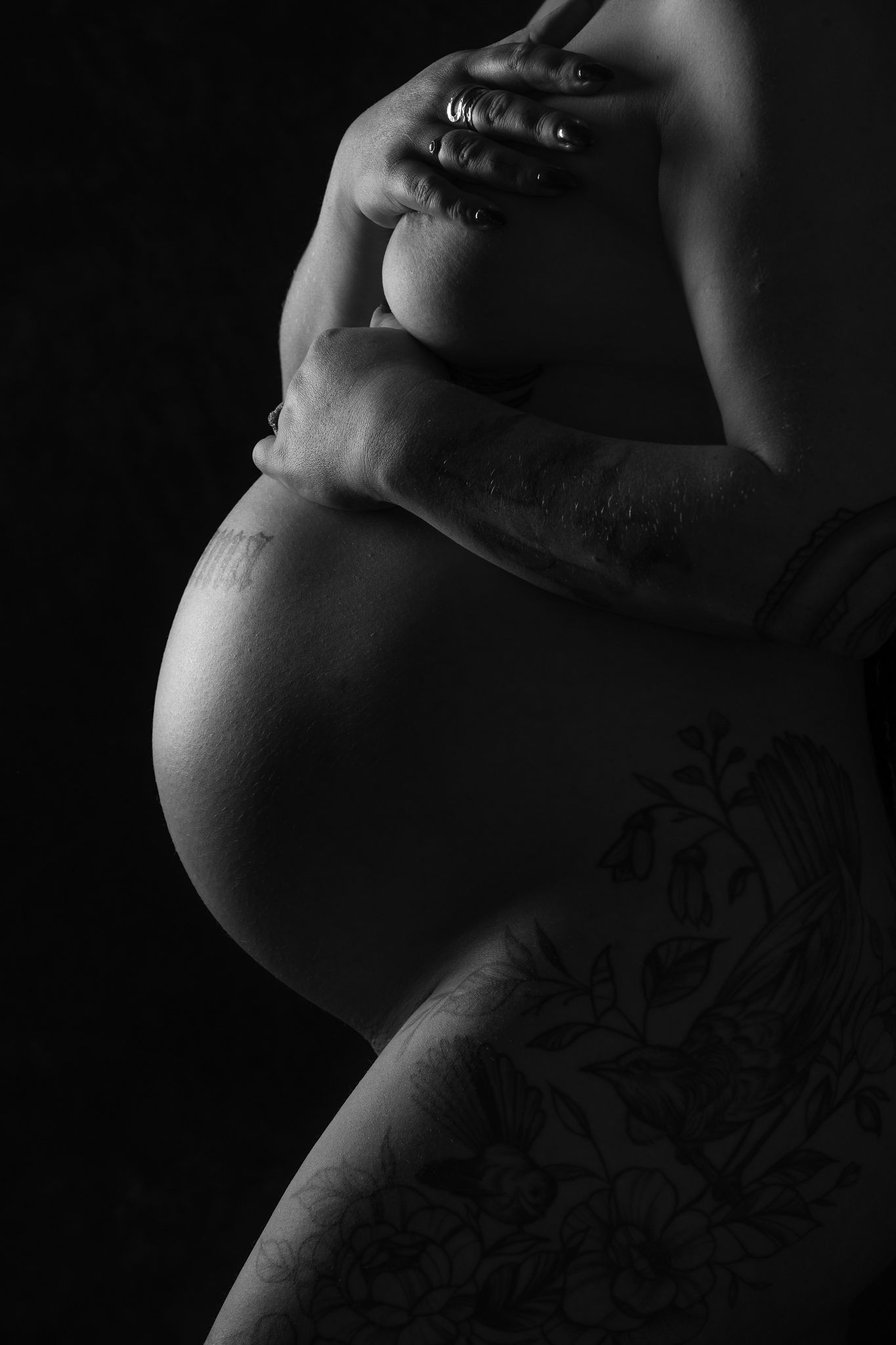Black and white maternity photo of pregnant woman cradling bare baby bump with tattooed arm against dark background.